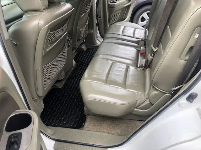 The backseat area of a car with leather seats, seat belts, and a black rubber floor mat.