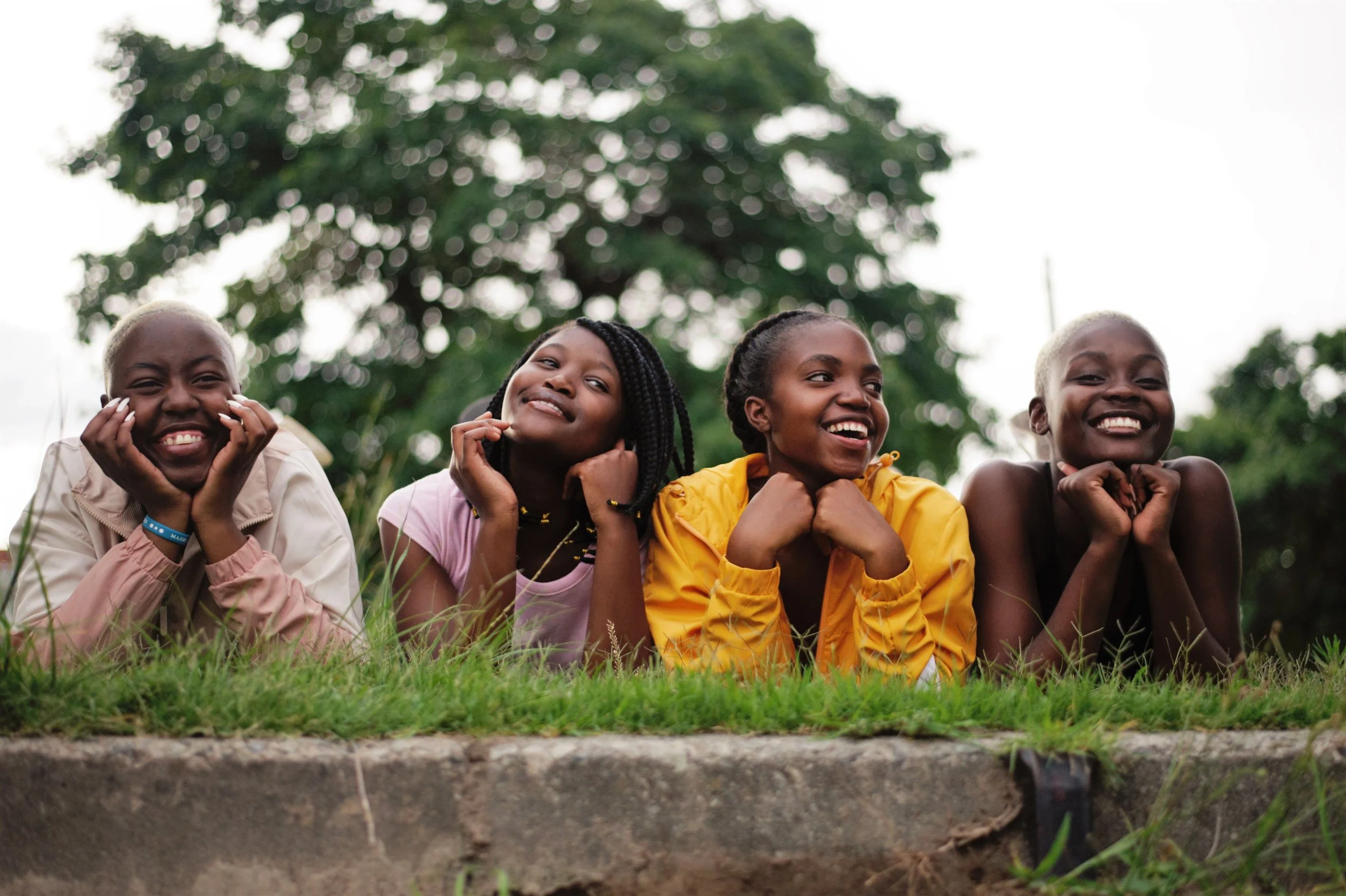 Four happy young girls with dark skin lying on the grass in a park, smiling and looking cheerful, with a large tree in the background and a bright sky.