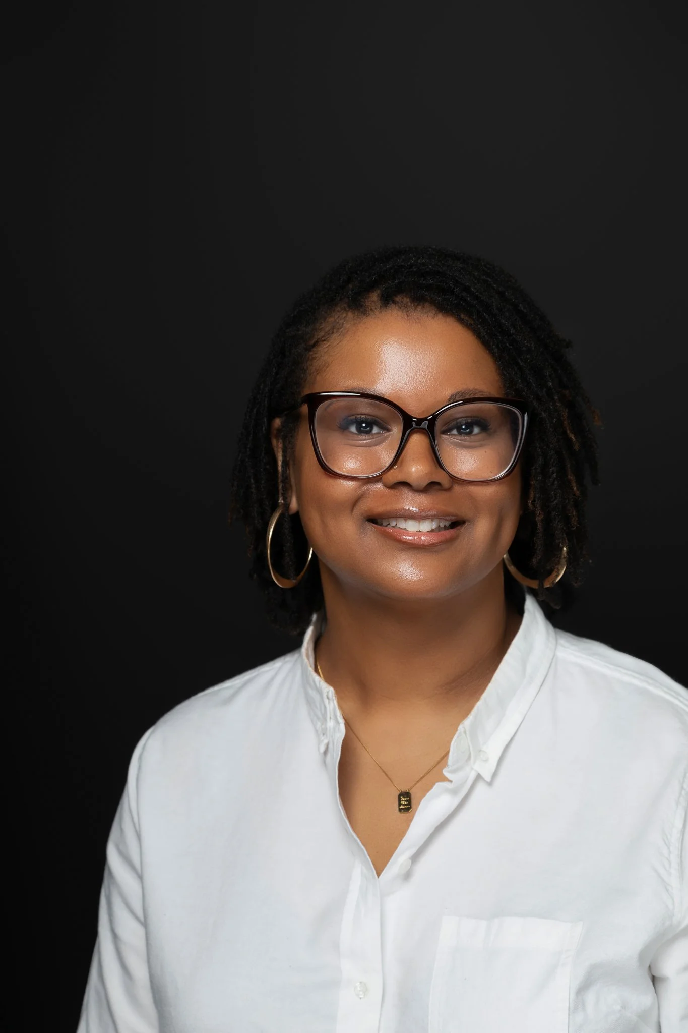 A Black woman with a caramel complexion and dark microlocs and black-framed glasses, smiling, wearing a white button-up shirt and a necklace, against a black background.