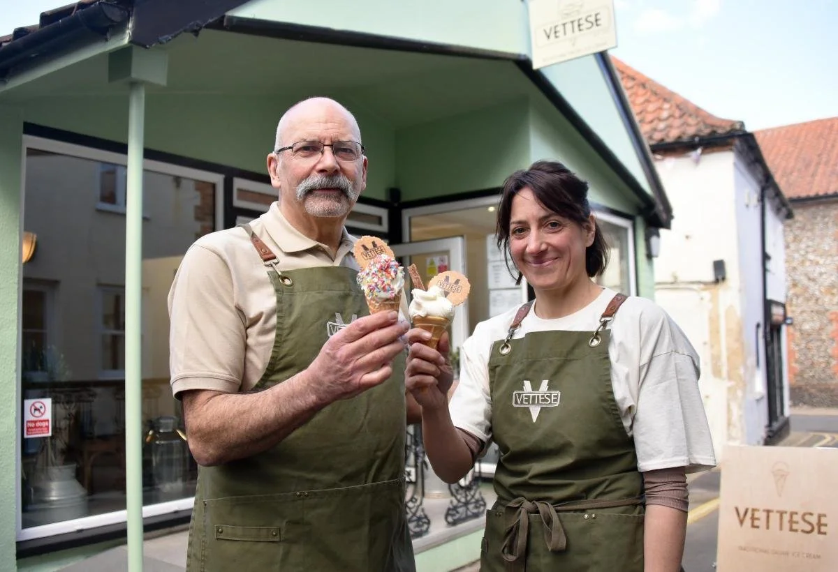 A man and woman standing outside an ice cream shop, each holding a cone of ice cream with colorful sprinkles. They are wearing aprons with the logo 'VETTESE' and smiling at the camera. The shop has a green exterior and a sign with 'VETTESE' on it.