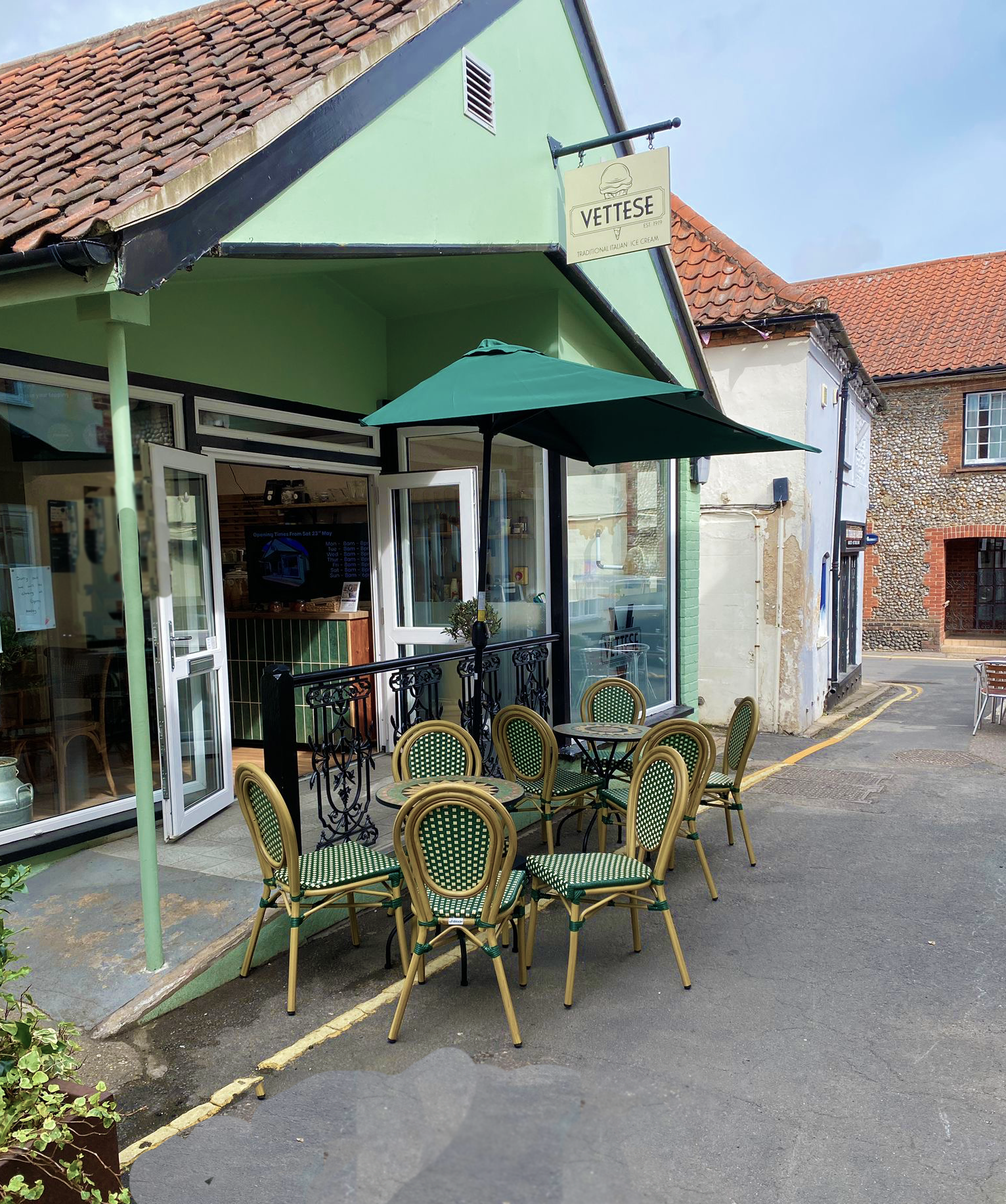 A cozy outdoor seating area with several green and yellow checkered chairs and small round tables, shaded by a green umbrella outside a restaurant or café called Vettese, featuring glass doors and a green exterior wall.