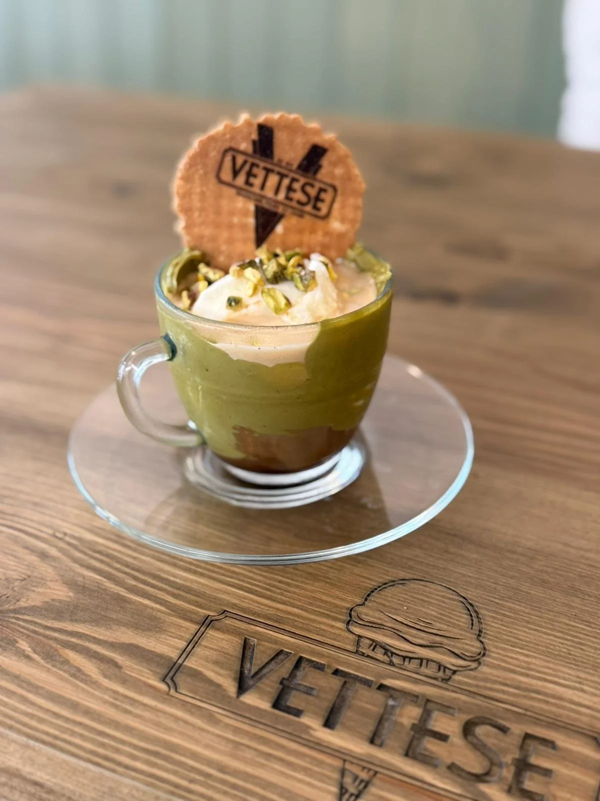 Small glass cup of pistachio-flavored dessert topped with whipped cream, chopped pistachios, and a cookie with the word 'VETTESE' printed on it, placed on a wooden tray with a logo.