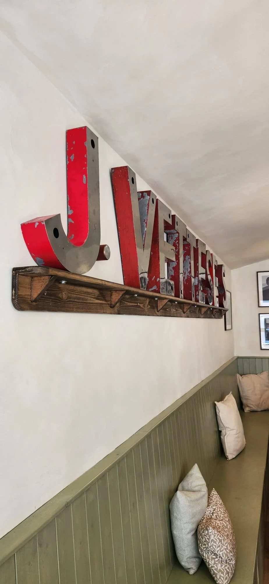 A decorative sign on a white wall spelling the word 'LOVE' with the letters made of metal and painted red, mounted on a wooden ledge; the sign shows signs of distress and weathering.