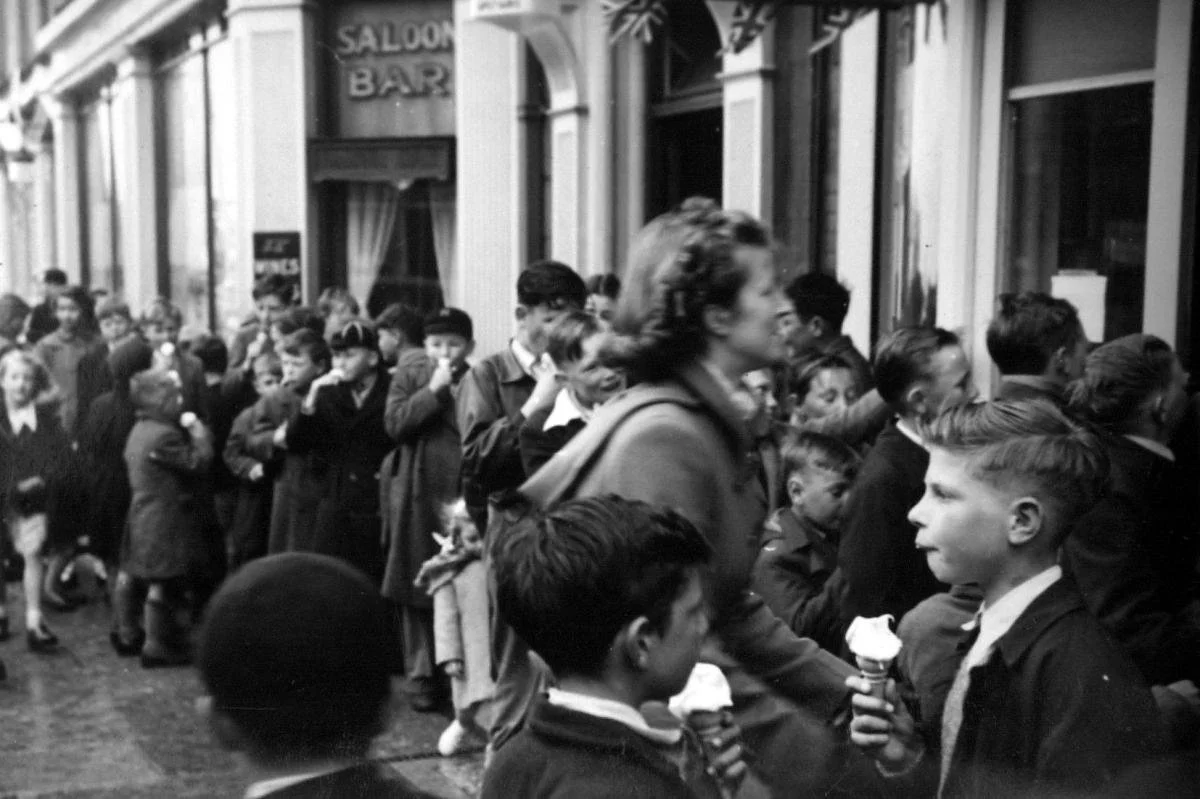 A black-and-white photo of a long line of children waiting outside a building with windows and a sign that reads 'Saloon Bar'. The children appear to be holding ice cream cones and are dressed in 1940s-style clothing.