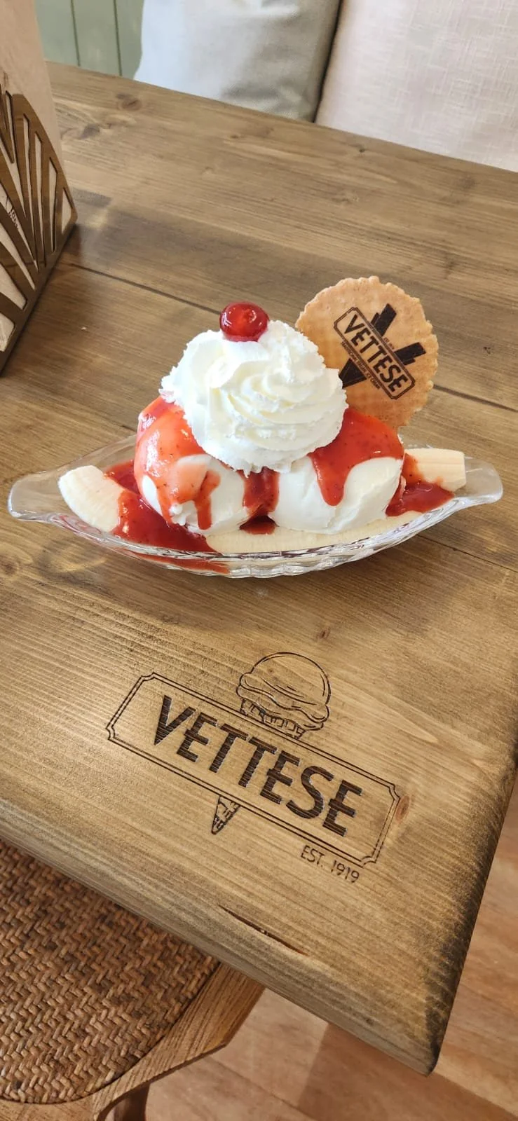 Banana split sundae with whipped cream, cherry, strawberry syrup, and a Vetise cookie in a glass dish on a wooden table with Vetise logo.