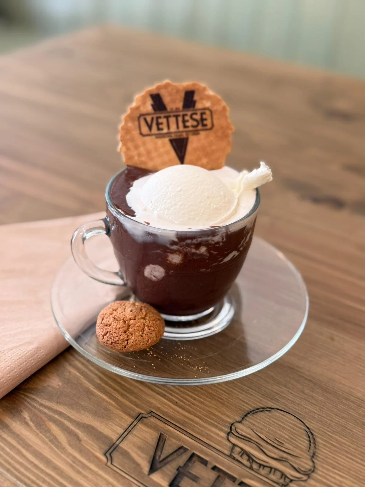 A glass mug of hot chocolate topped with vanilla ice cream, garnished with a Vetese cookie. A small cookie rests on the saucer. The mug is on a wooden tray with Vetese branding.