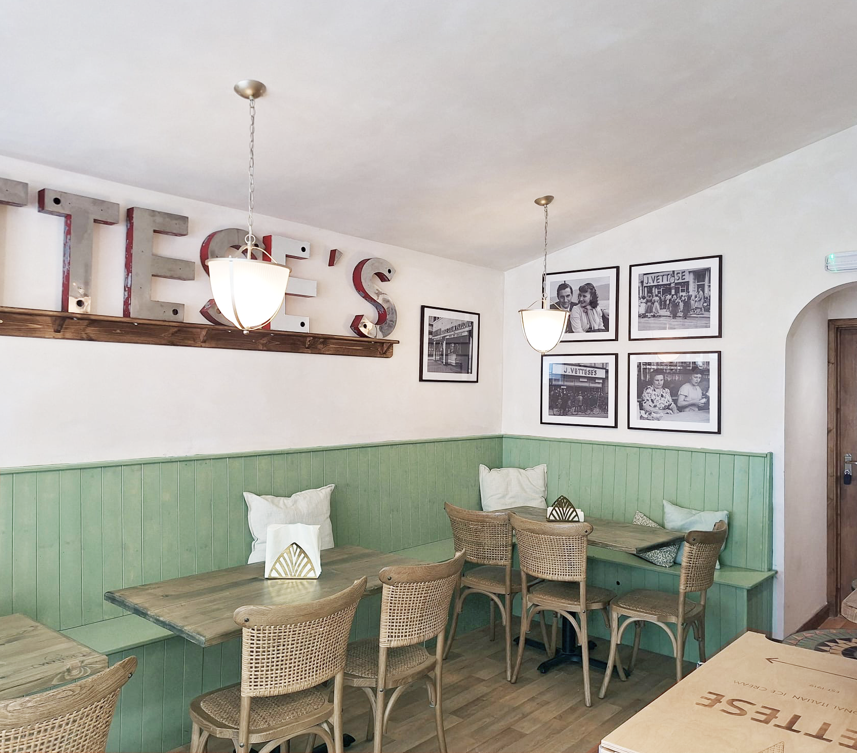 Interior of a cafe or restaurant with light-colored walls, green wainscoting, wooden tables and wicker chairs. Decorative black and white photographs on the walls and large metal letters spelling 'TEVS' on a shelf with hanging pendant lights.