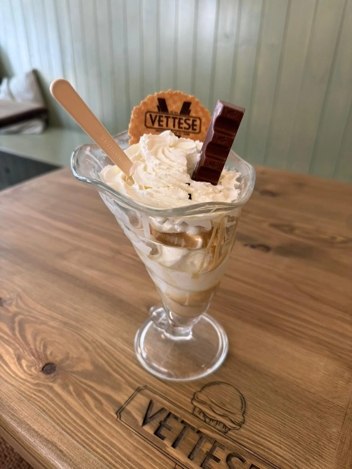 A glass of ice cream topped with whipped cream, a chocolate bar, a biscuit with "VETTESE" written on it, and a white spoon, on a wooden table with a "VETTESE" logo engraved.