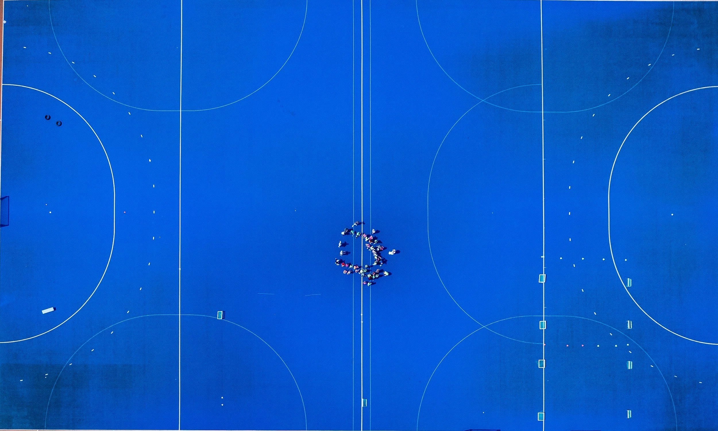 An aerial view of a large blue sports court with a group of people forming a heart shape in the center and scattered sports equipment on the court.