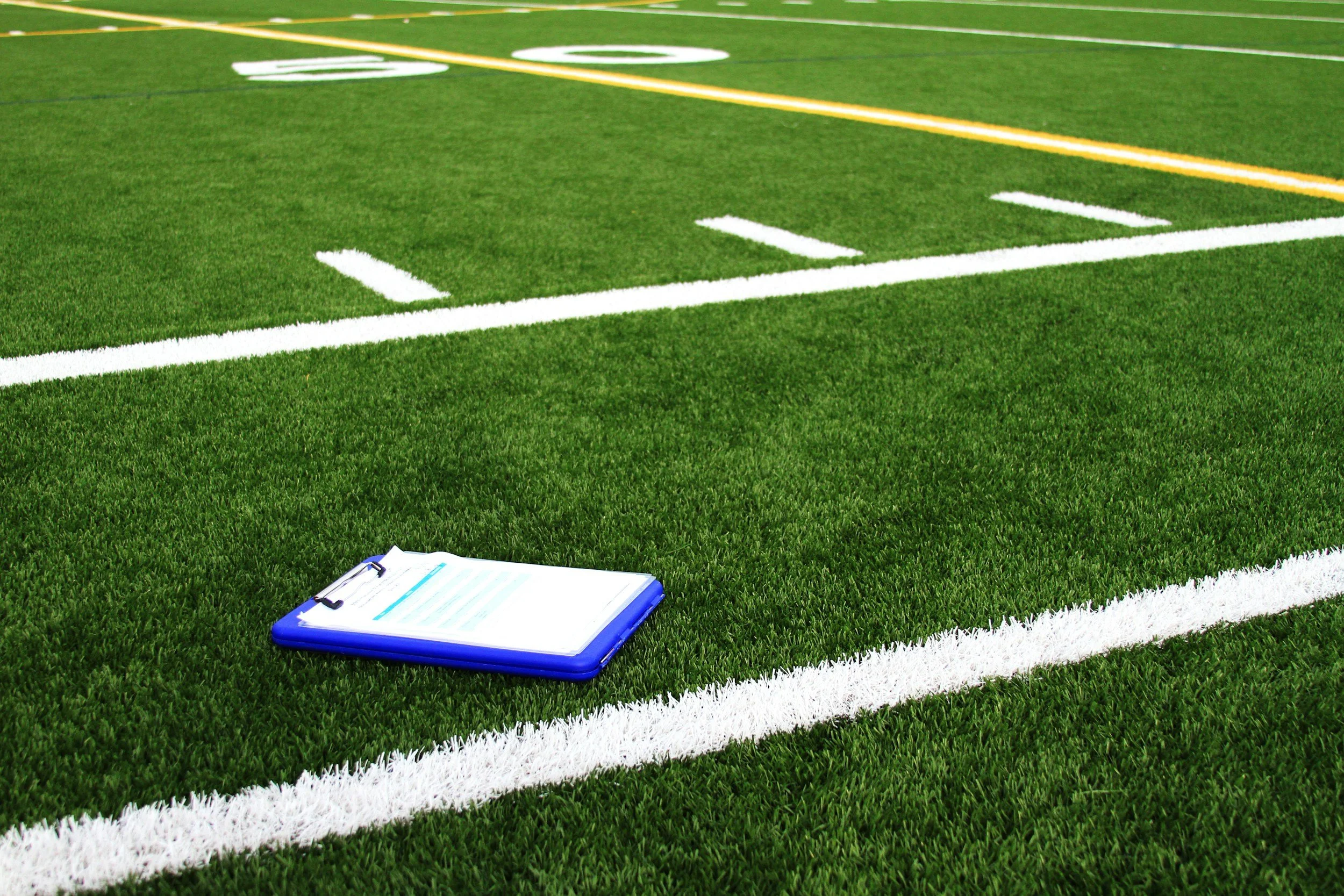 A section of a green football field with white yard lines and a yellow line marker. A clipboard with papers is on the grass near the bottom of the image.