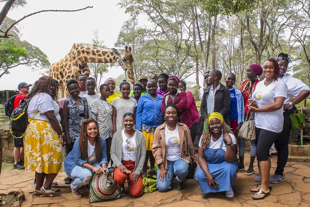 Group of people, including children and adults, posing outdoors with a giraffe in the background, trees, and cloudy sky.
