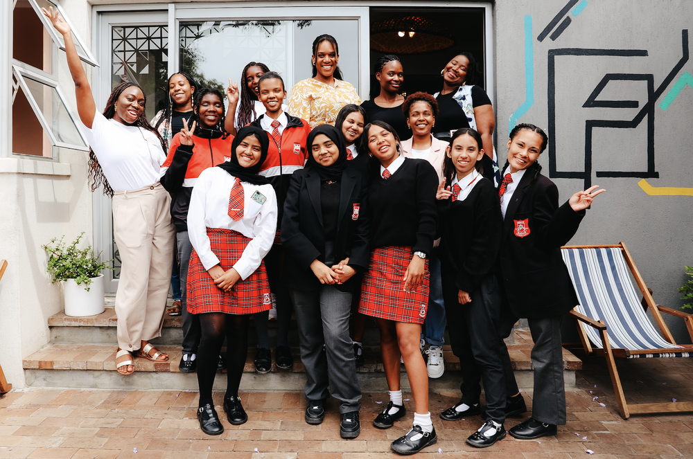 Group of young girls and women smiling and posing together outside a building, some making peace signs, in school uniforms and casual clothes.