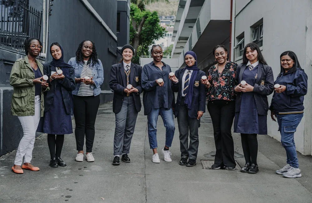 Nine women standing outdoors in an alleyway holding cups, smiling, some wearing school uniforms and jackets.