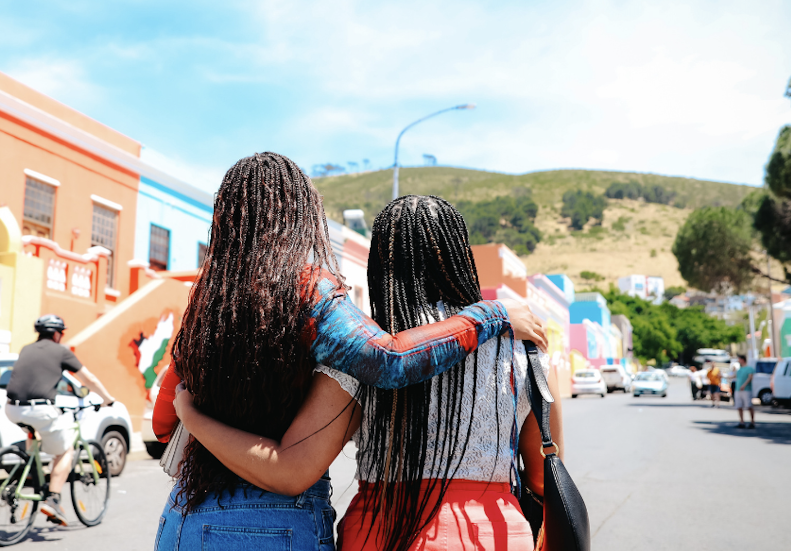 Two women with long braided hair are walking together with their arms around each other in a colorful town, with hills and a blue sky in the background.