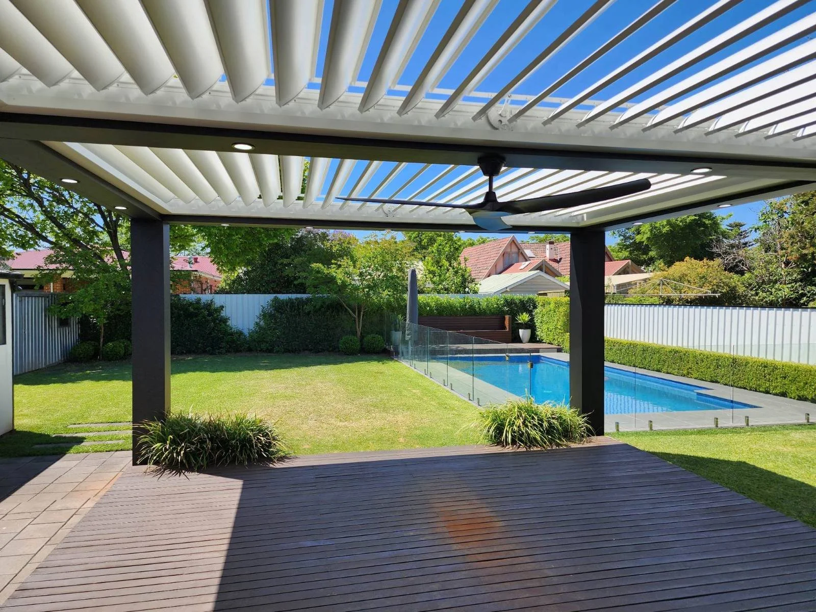 View of backyard patio with wooden decking, that overlooks a green lawn and swimming pool enclosed by a glass fence, with trees and rooftops in the background.