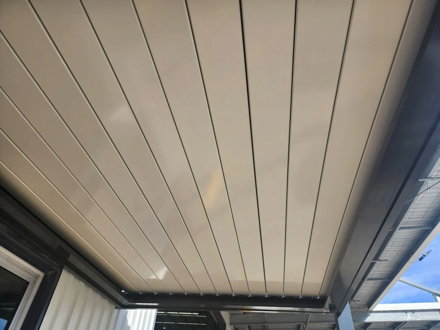 View of a beige, paneled ceiling of an outdoor patio or balcony area with a metal railing visible at the edge and a partly cloudy sky.