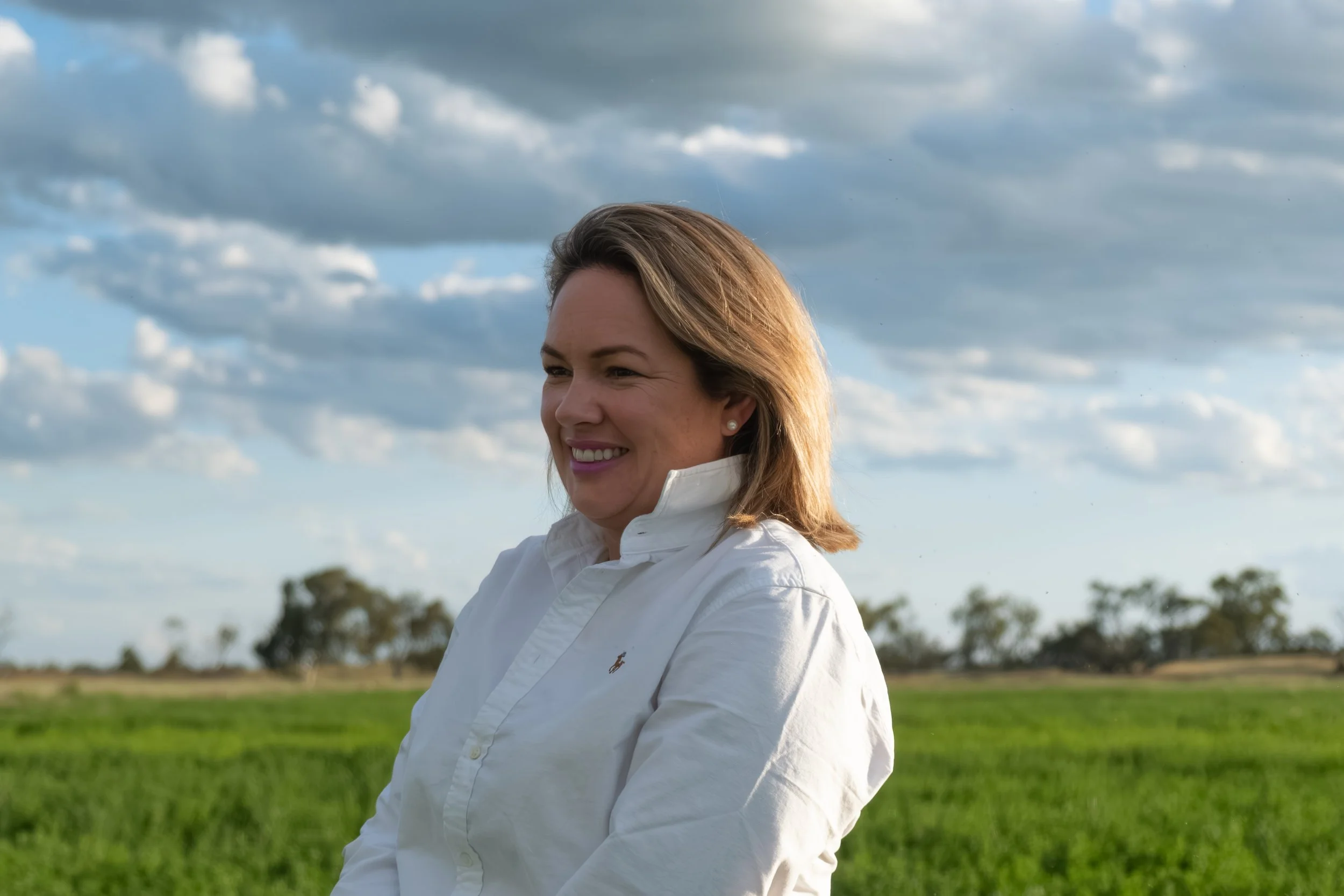 A woman smiling outdoors with a partly cloudy sky and a green field in the background.