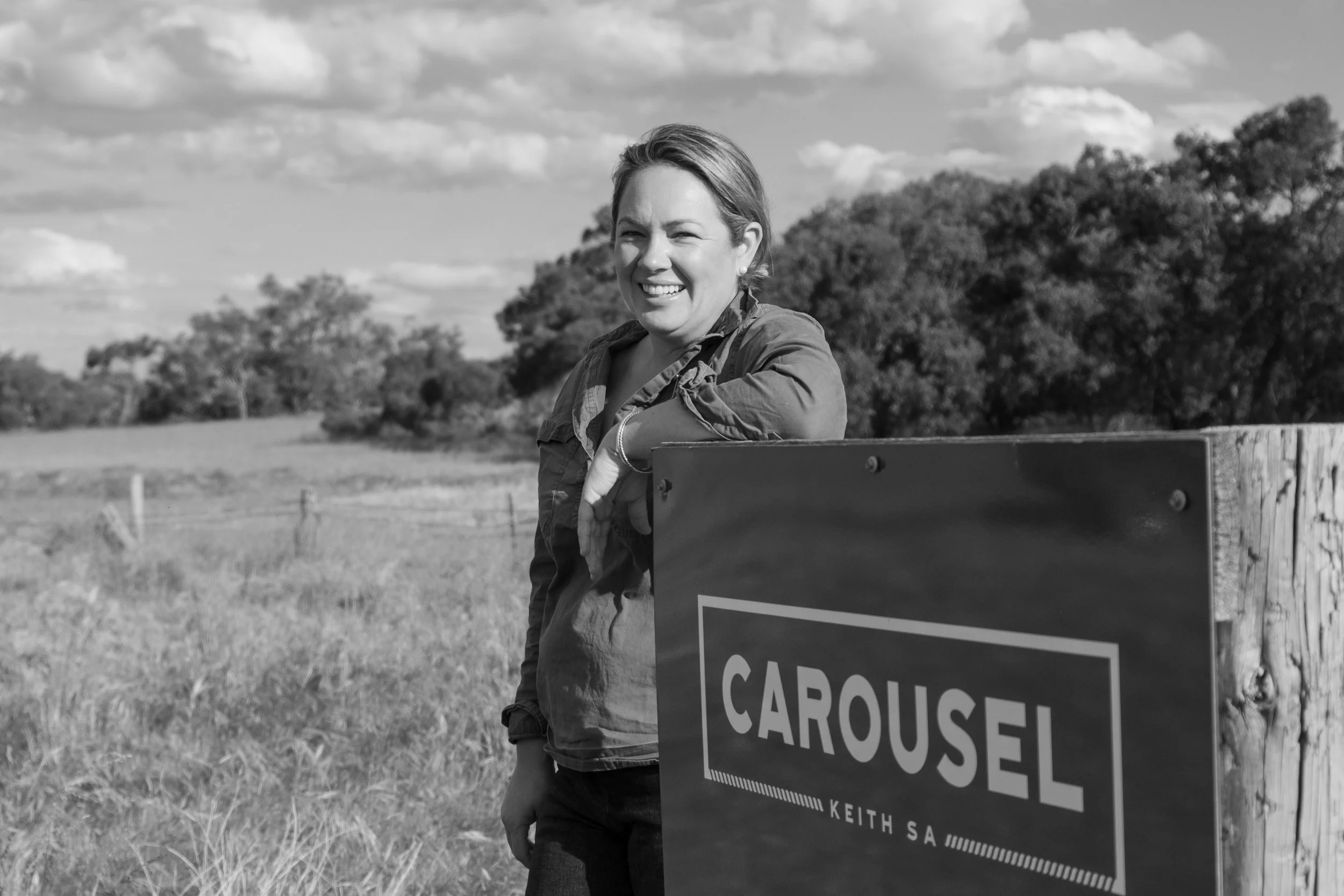 A woman with short hair smiling outdoors near a fence, holding a large sign that reads 'Carousell' in a rural setting with trees and a cloudy sky in the background.