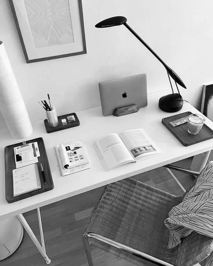 A minimalist white desk with a MacBook, open books, notebooks, pens, a candle, a small tray with stationery, a desk lamp, and a glass of water, with a striped chair and a picture frame on the wall.