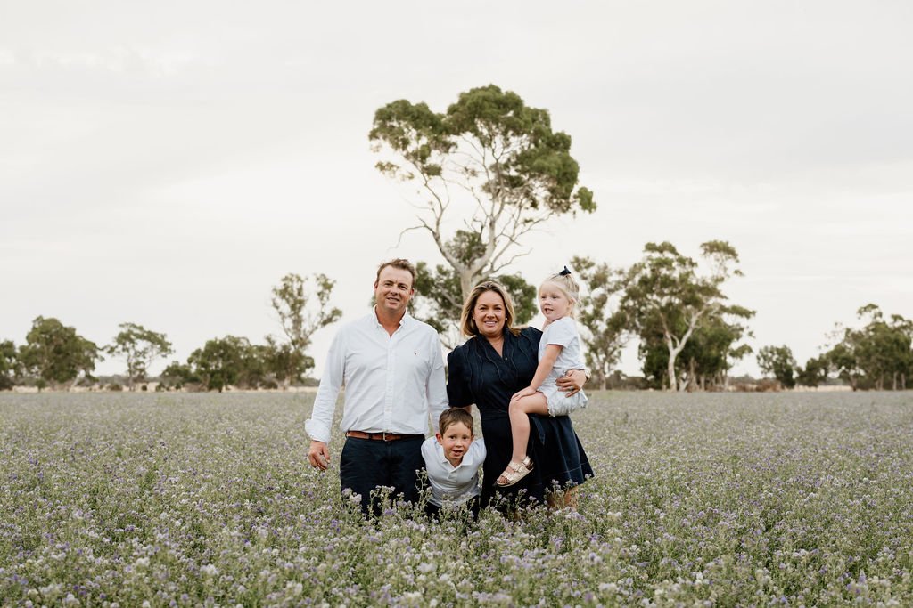 A family of four stands in a lavender field with trees in the background, smiling at the camera.
