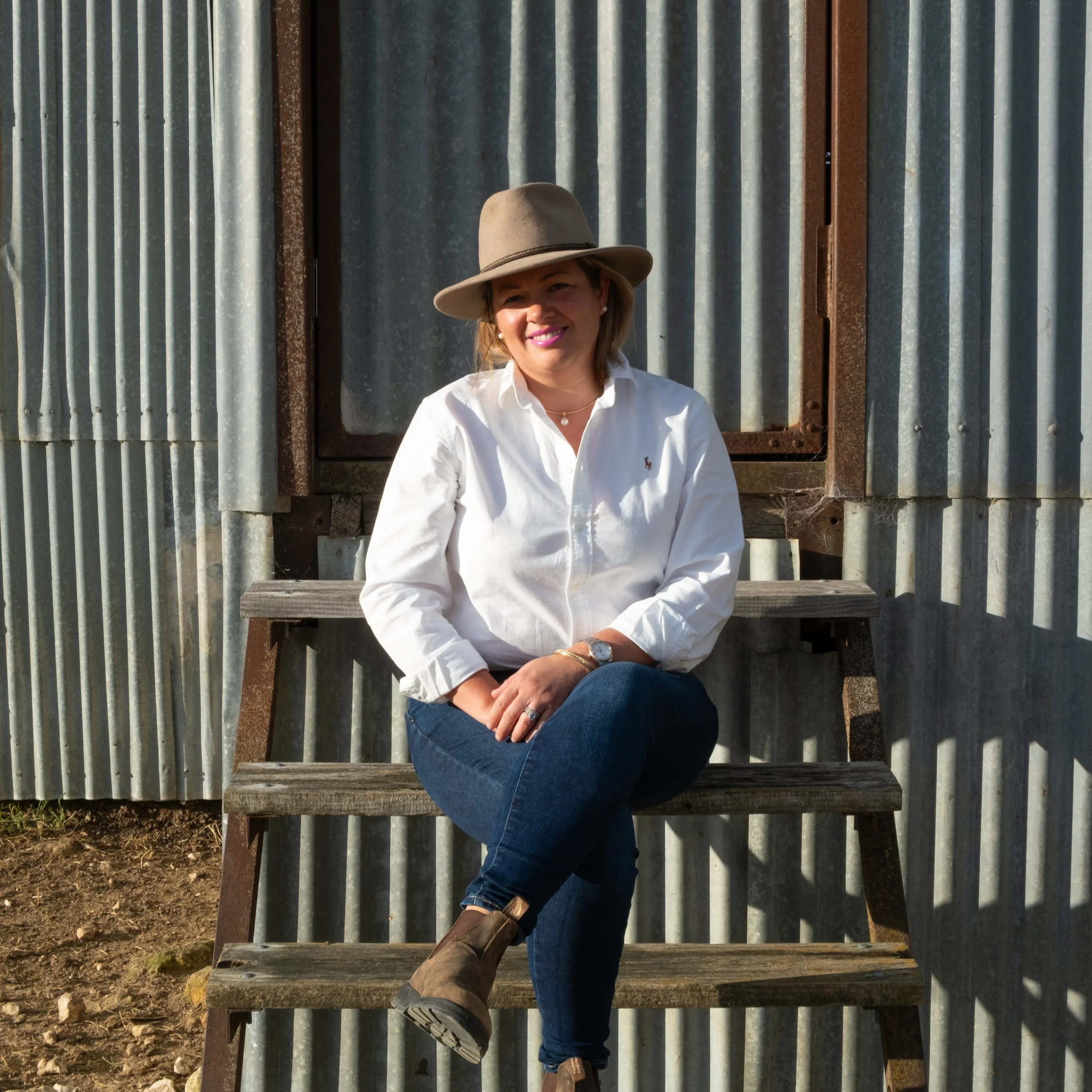 A woman with a tan hat, white blouse, and blue jeans sitting on rustic wooden benches in front of a weathered corrugated metal wall, smiling and looking at the camera.