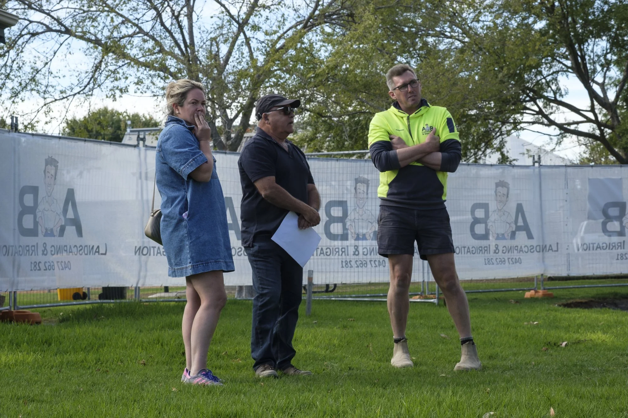 Three people standing outdoors on a grassy area, observing something. One woman wearing a denim jacket and sneakers on the left, a man in the middle with sunglasses and a dark shirt, and a man on the right with glasses, a bright yellow jacket, and shorts. In the background, there is a white fence with printed text and illustrations of a cartoon character, and trees with green leaves.