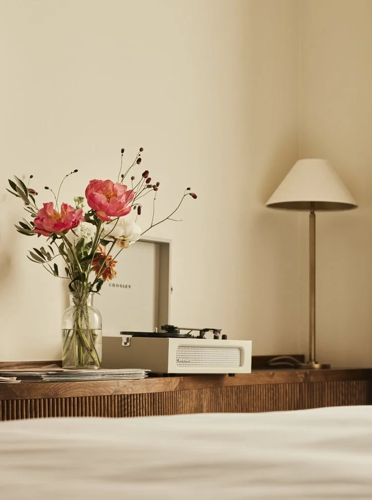 A bedroom scene with a wooden nightstand holding a glass vase of colorful flowers, a vintage record player, and stacked magazines. A white table lamp is on the right, and a framed photo or artwork is behind the flowers.