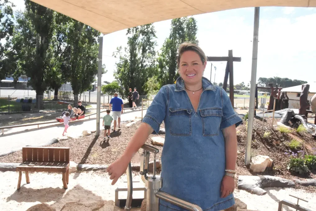 A woman smiling outdoors in a park or playground area with trees and children in the background.