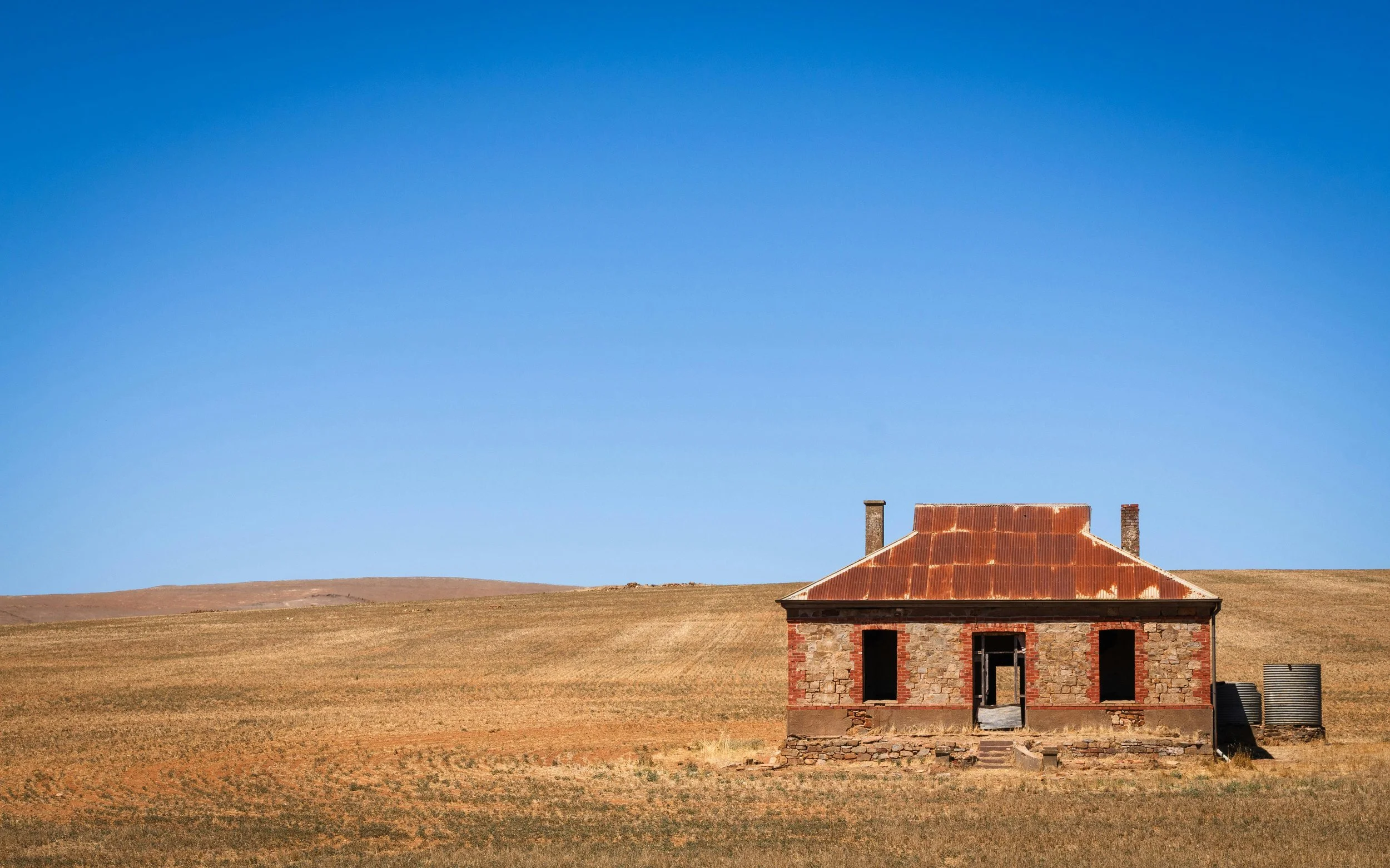 An old, partially built stone house with a rusted metal roof, standing alone in a vast, flat, grassy landscape with a clear blue sky in the background.