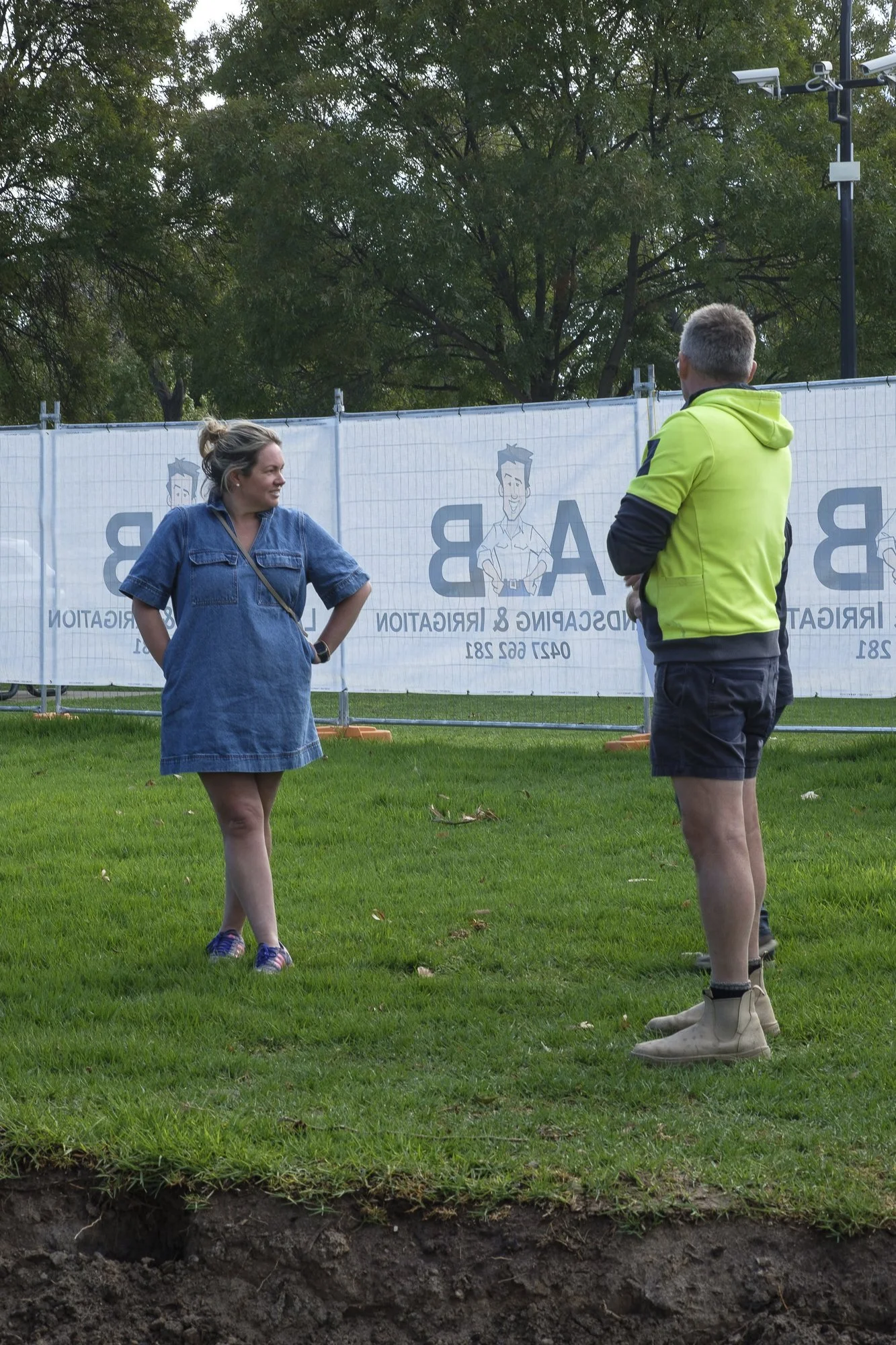 A woman and a man stand on a grassy area, engaged in conversation. The woman is wearing a denim dress and sneakers, with her hands on her hips. The man is dressed in a bright yellow jacket, black shorts, and beige boots, seen from the back. In the background, there is a fence with signage and trees. The foreground shows a cross-section of earth and soil.