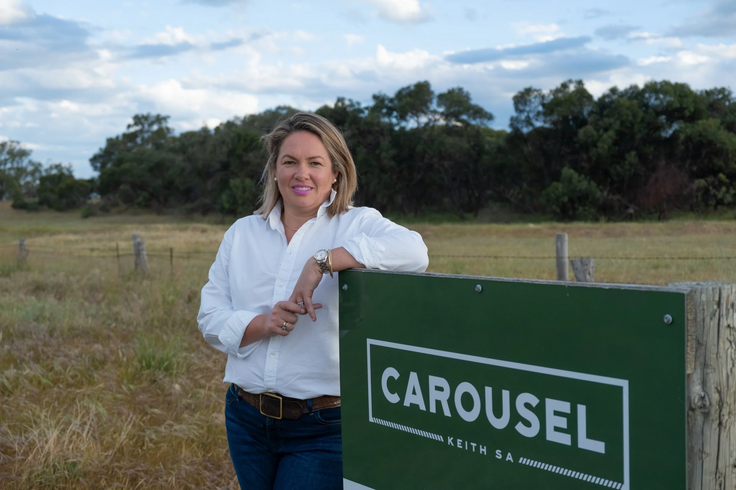 Woman in white shirt leaning on a green sign with white text outdoors with trees and a partly cloudy sky in the background.