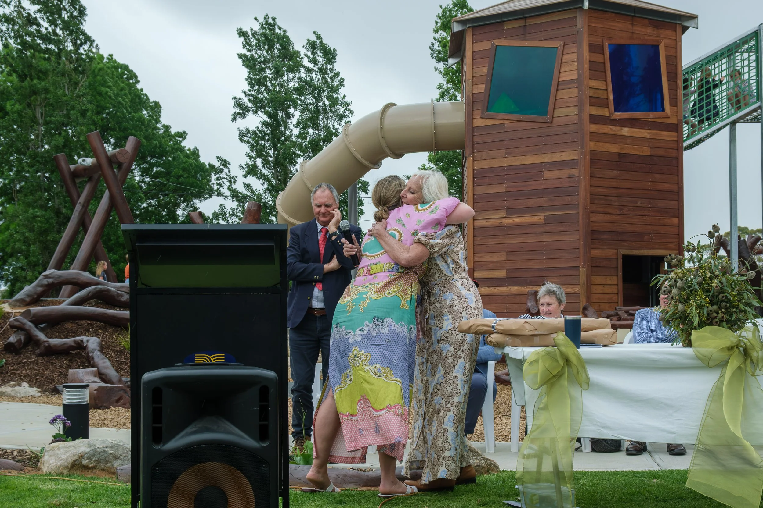People hugging at an outdoor gathering near a wooden play structure with a slide, surrounded by trees and a table with a yellow tablecloth.