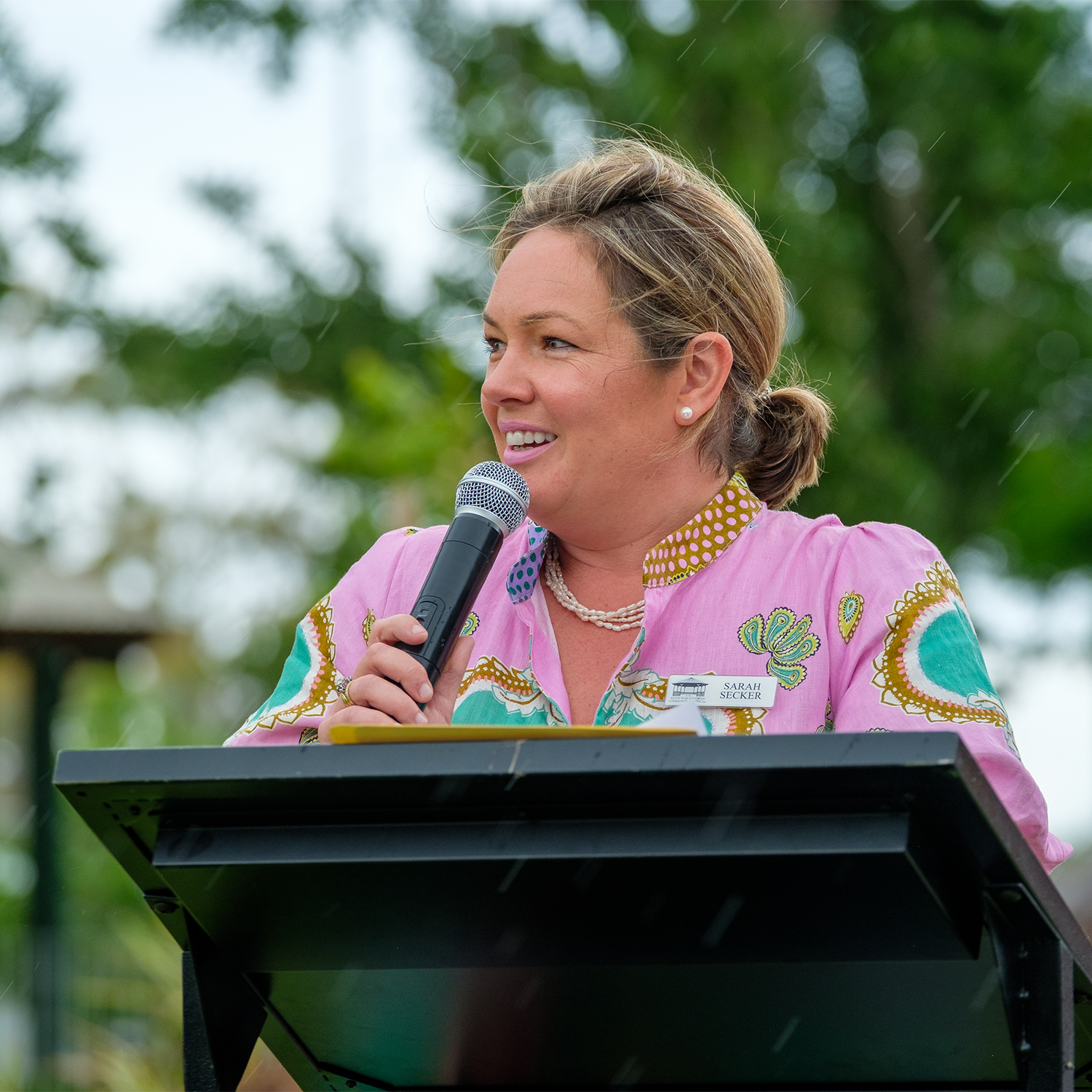 A woman speaking into a microphone at a podium outdoors with trees in the background.