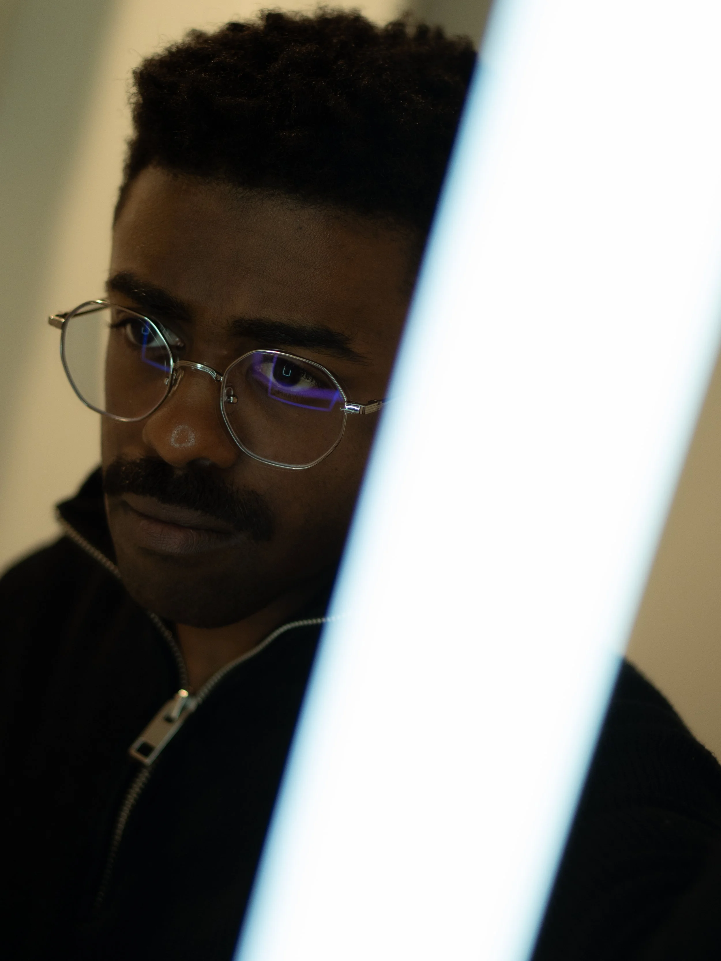 A person with dark curly hair, wearing glasses, dressed in a dark hoodie, is looking at a bright electronic screen.