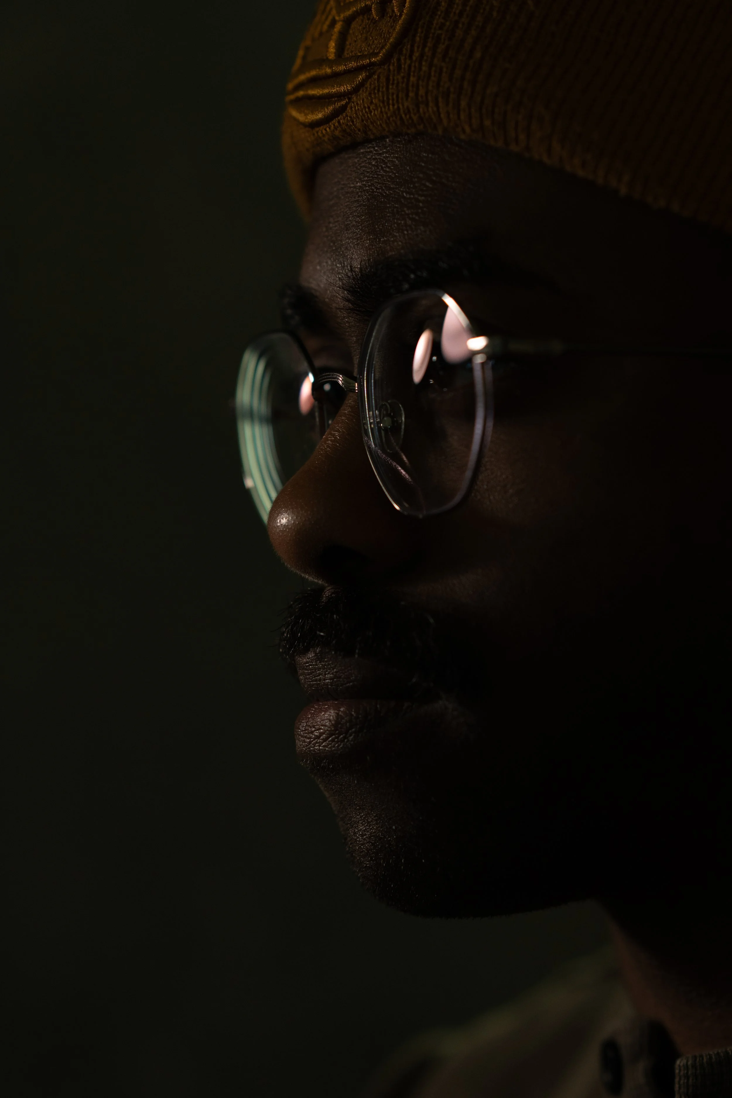 A close-up side profile of a person with dark skin, wearing glasses and a brown hat, against a dark background.