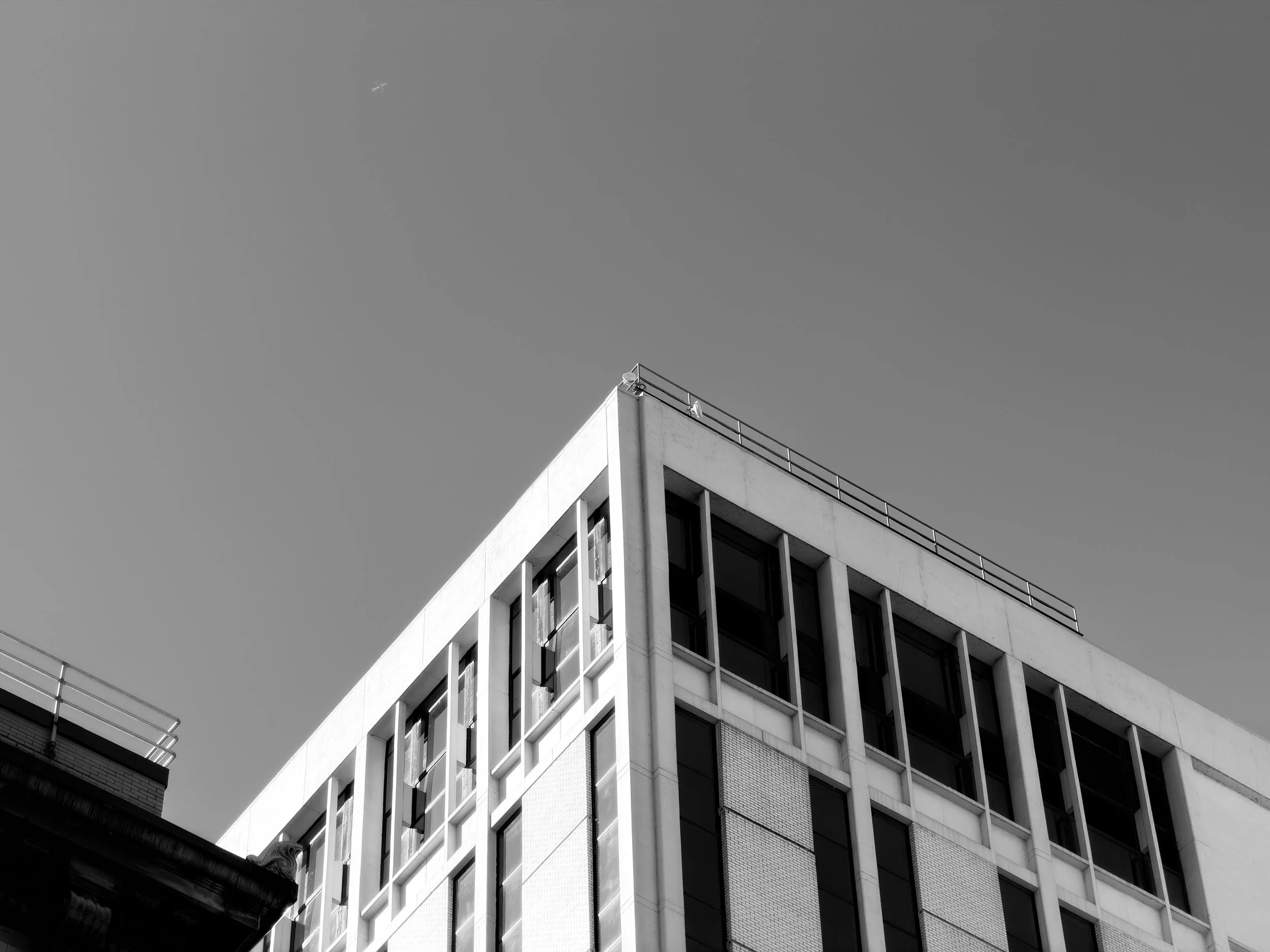 Black and white photo of a modern multi-story building with a corner view, featuring windows and antennas on the roof, against a clear sky with a small airplane in the distance.