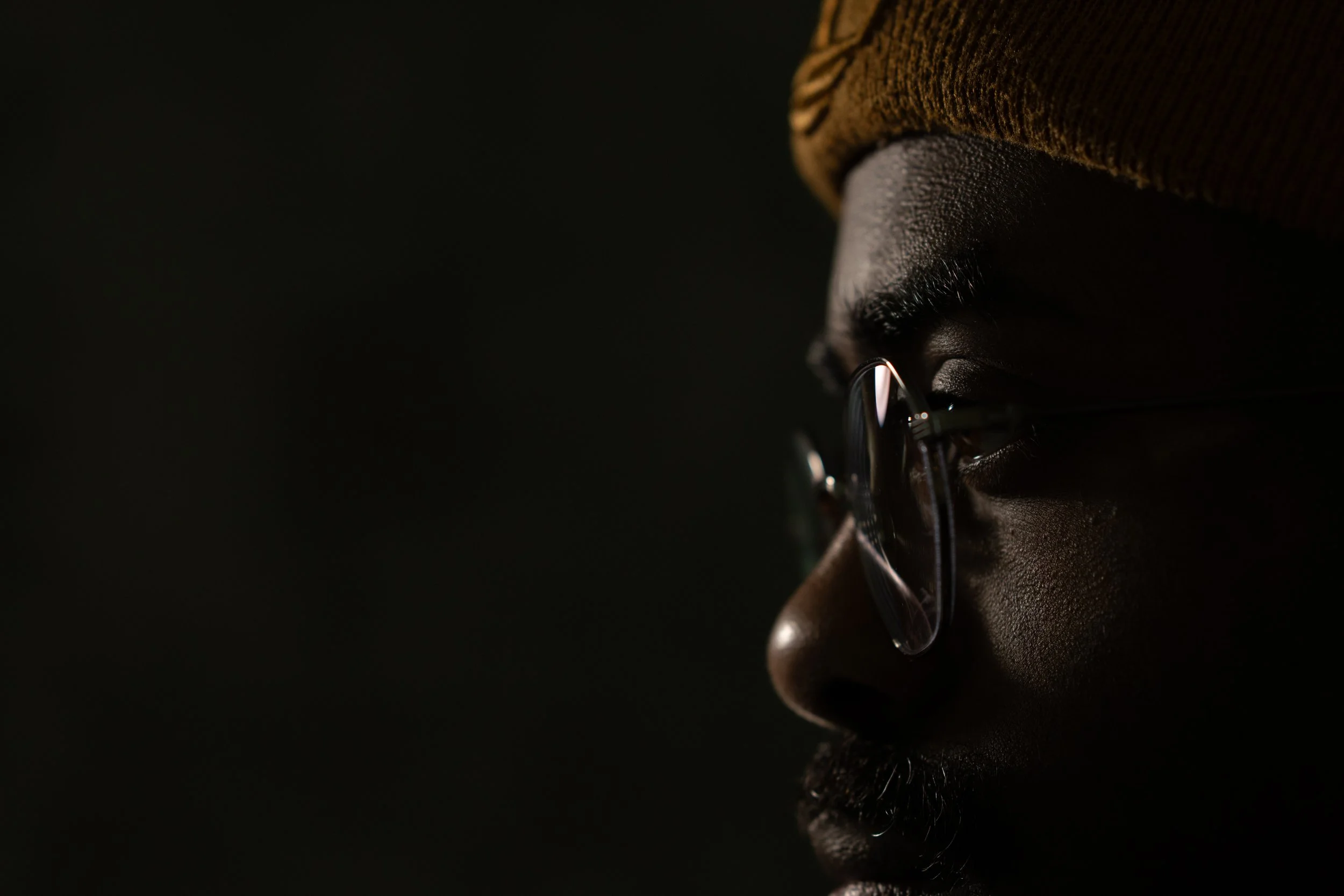 Close-up of a person wearing glasses and a brown knit cap, seen from the side in low lighting.