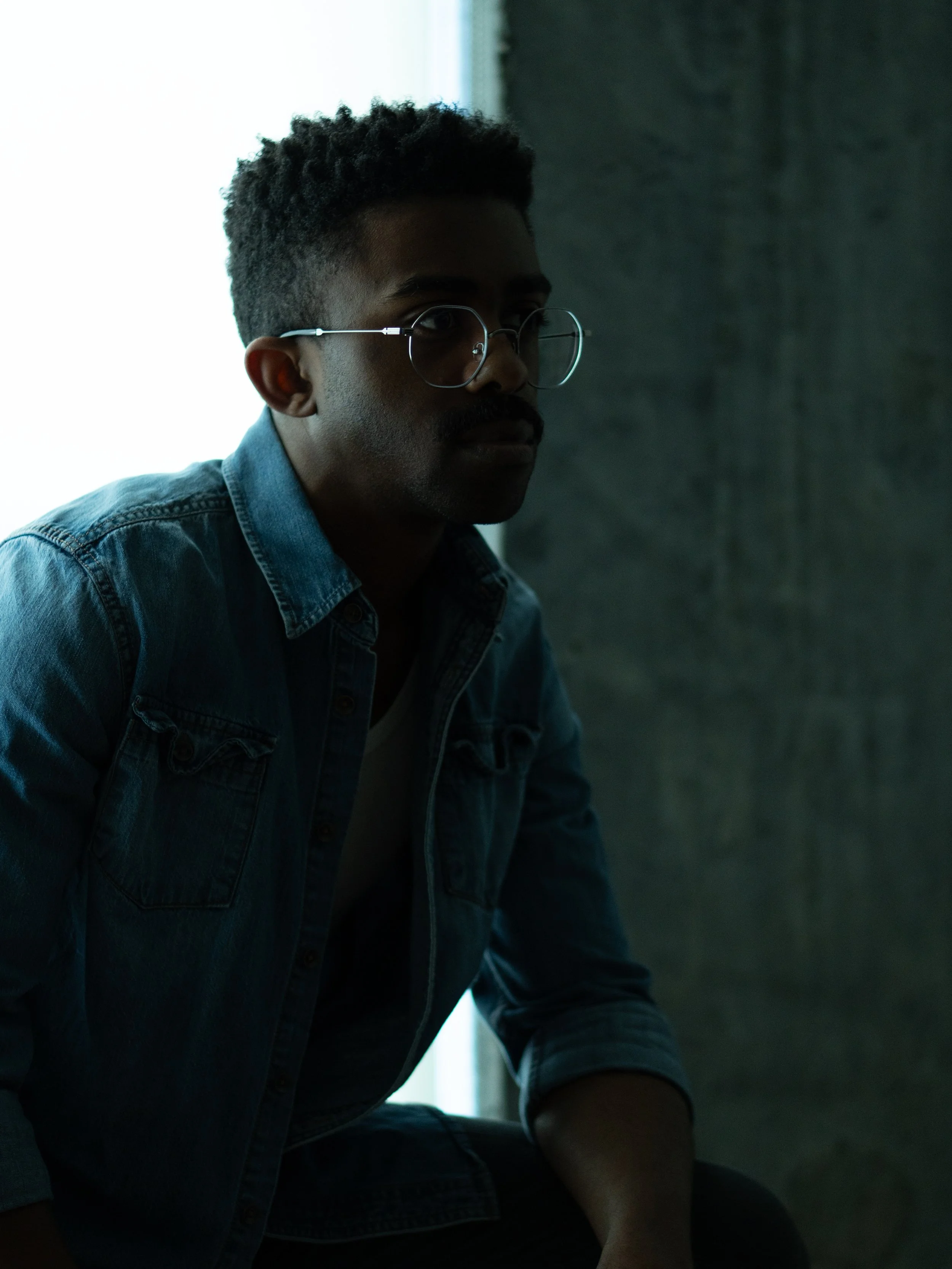 A young man with curly hair wearing glasses and a denim jacket, sitting in a dimly lit room near a window with light streaming in from behind.