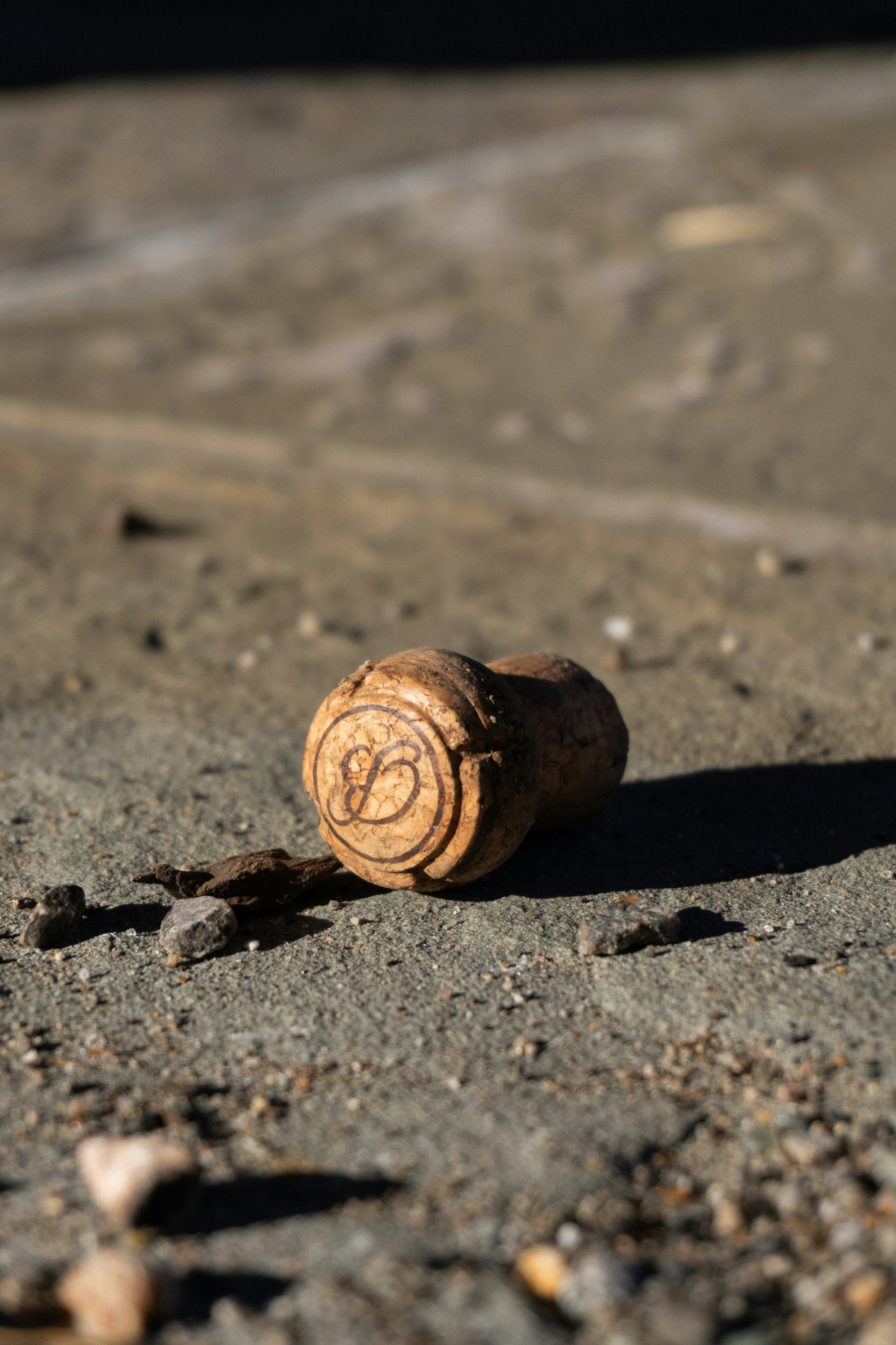 A cork golf ball marker with the letter B engraved on it, lying on sandy ground with small rocks and pebbles.