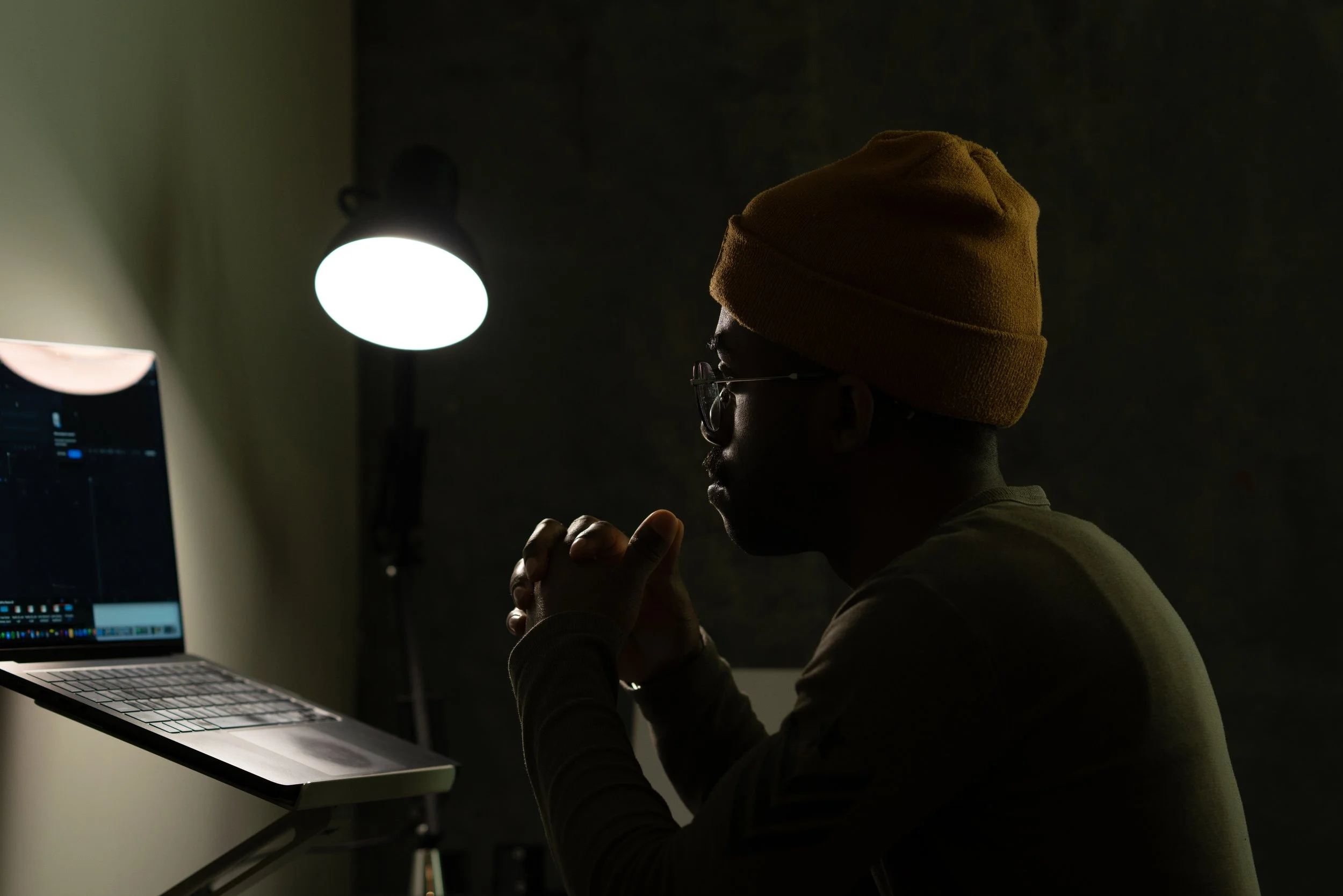 A person wearing a yellow beanie and glasses working in a dark room, illuminated by a computer screen and a desk lamp.