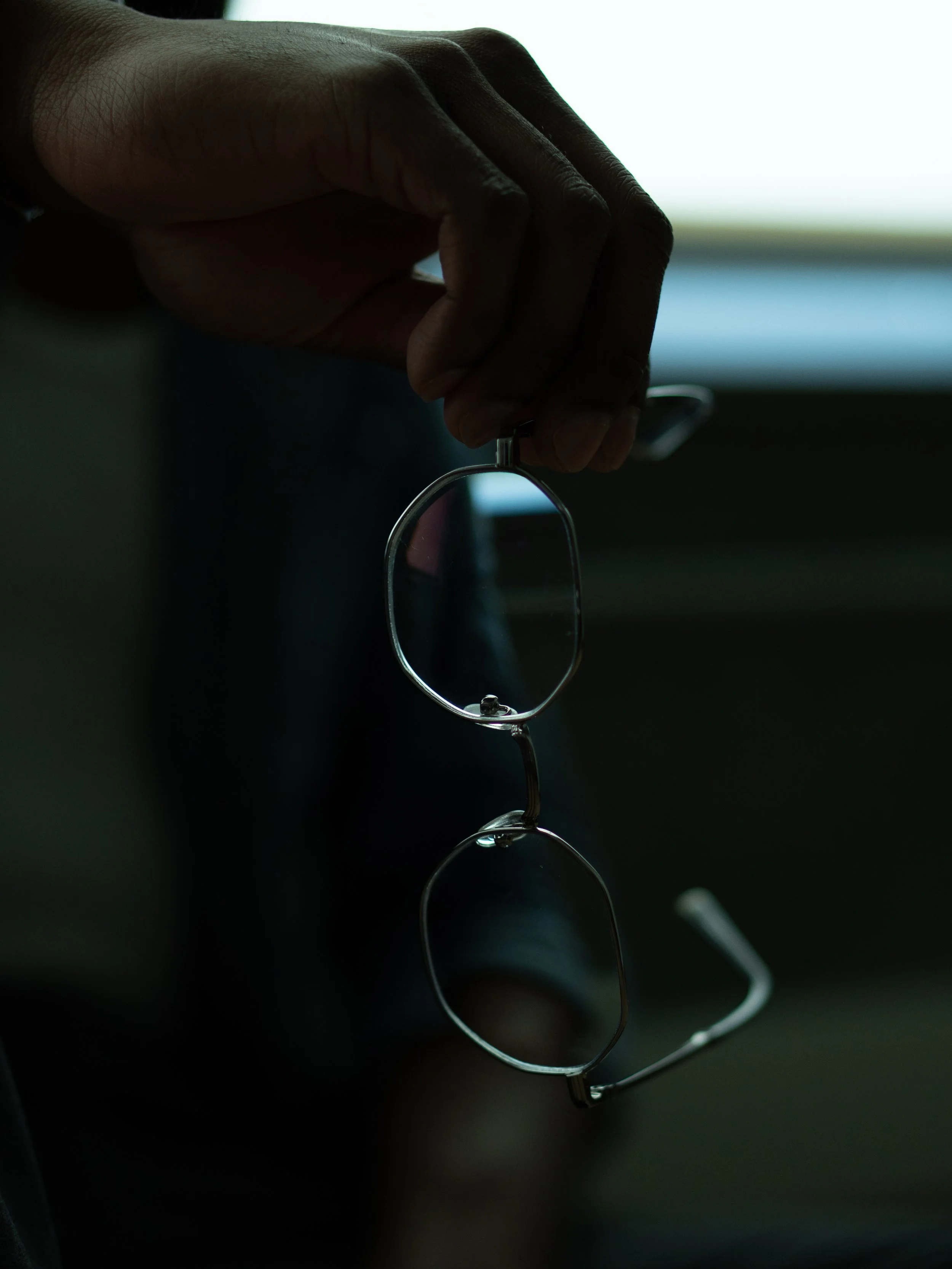A person's hand holding a pair of eyeglasses with a dark background and bright light behind.