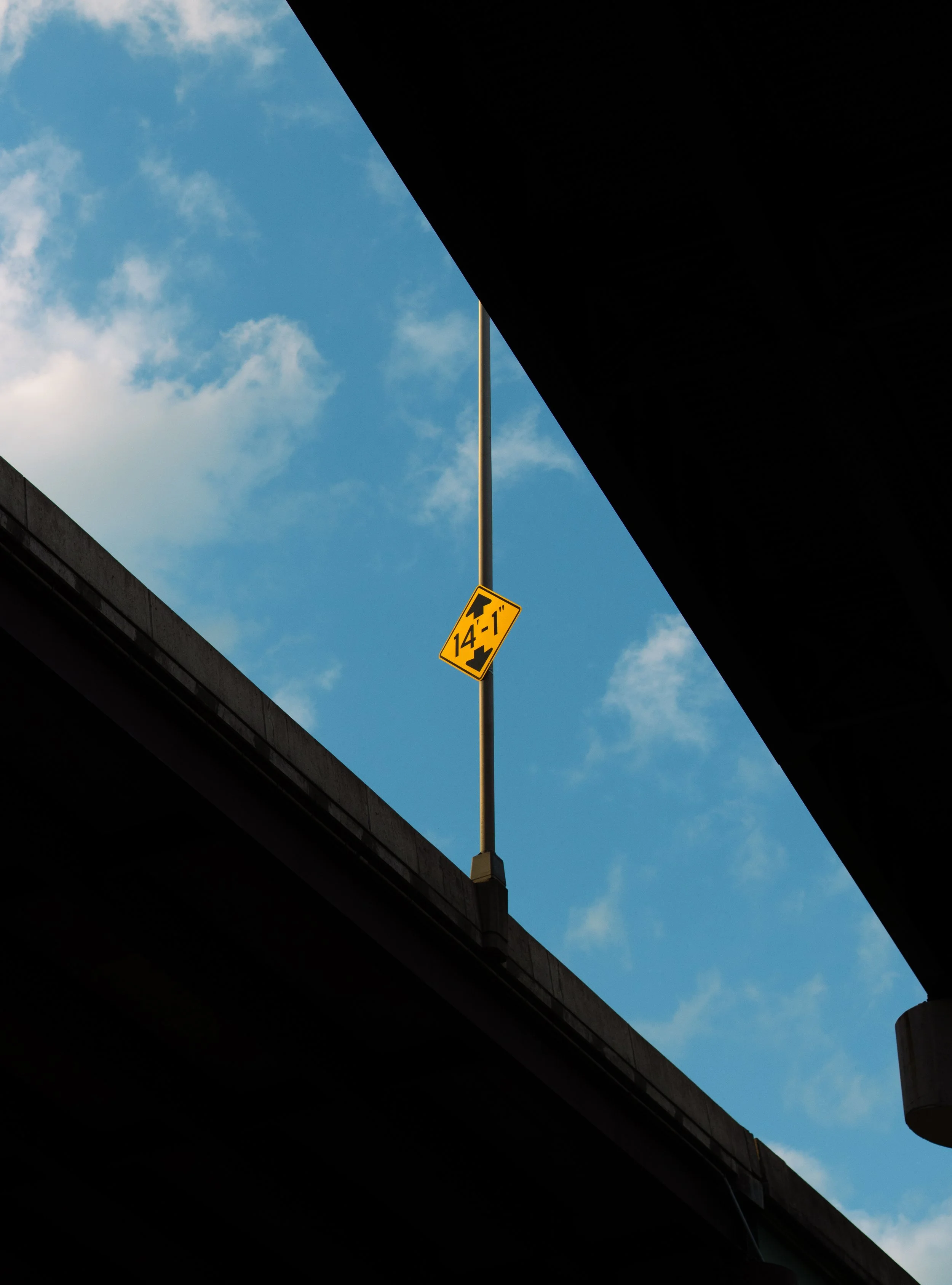 Photo showing the underside of a highway overpass with a yellow height clearance sign reading '14-1'. The sky is blue with some clouds, and the photo is taken from below, looking up through the space between two overpasses.