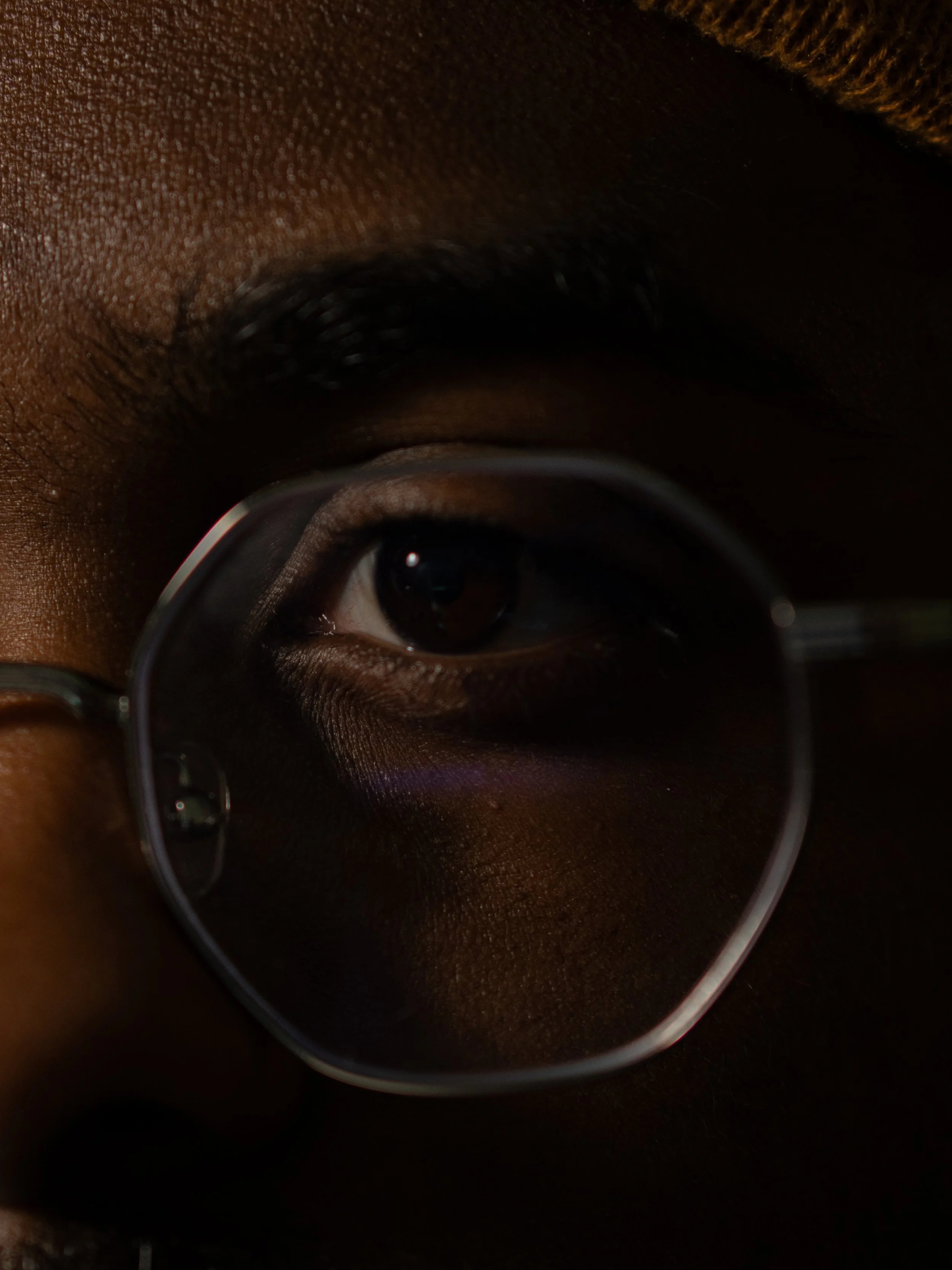 Close-up of a person's eye with dark skin, wearing glasses, reflecting the surrounding environment.