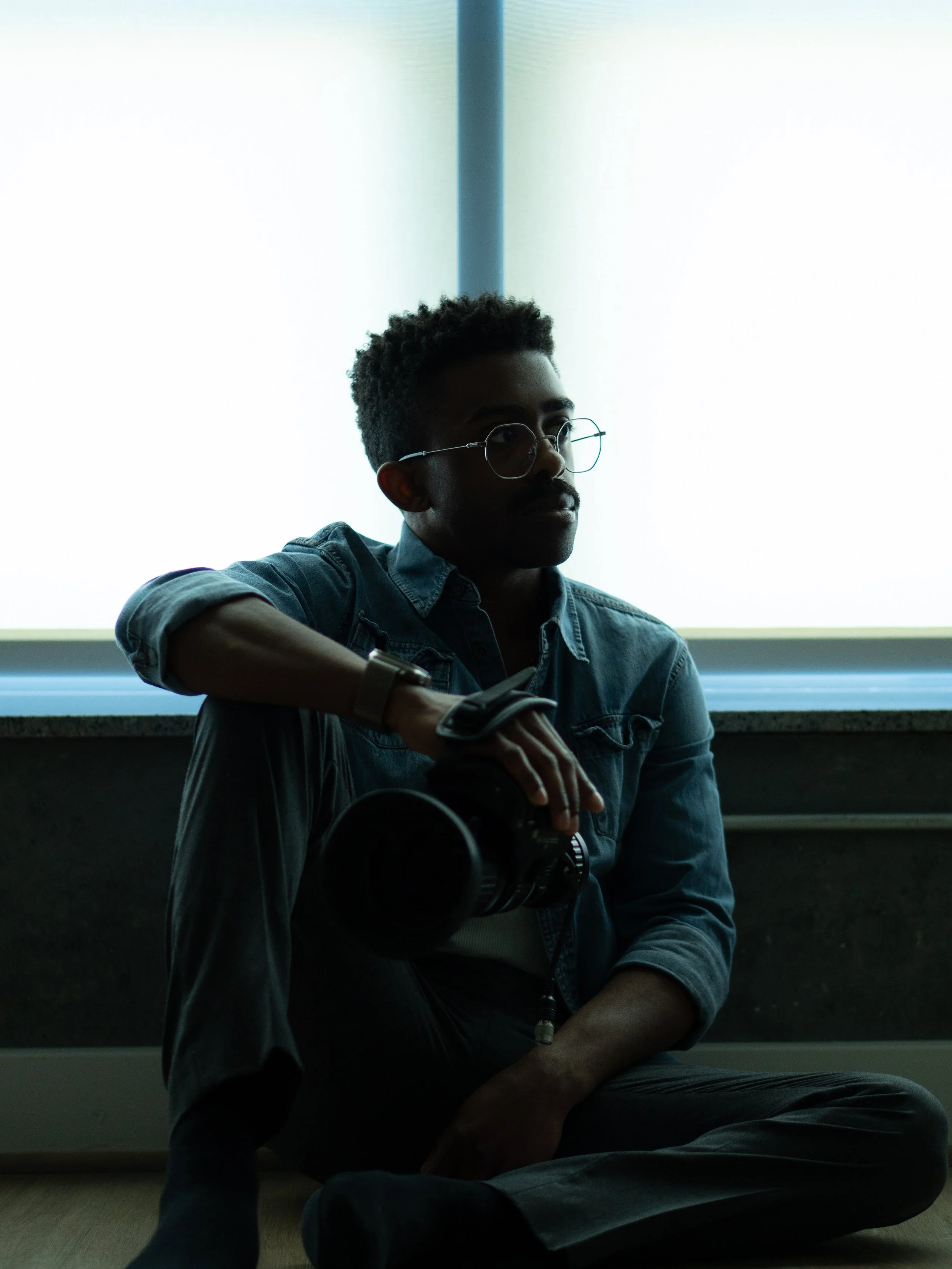 A man with short curly hair, glasses, and a mustache sitting on a windowsill with a camera in his lap, wearing a denim shirt, looking to the side, indoors.