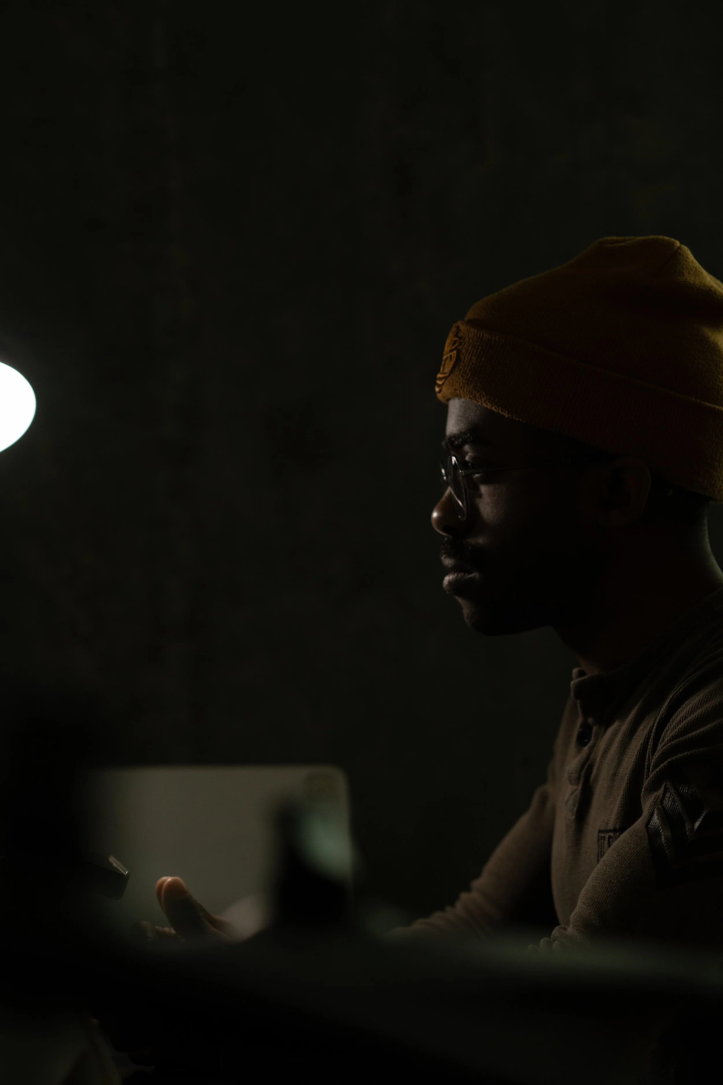 A side profile of a man wearing glasses and a yellow beanie, sitting in a dark room, looking at his phone.