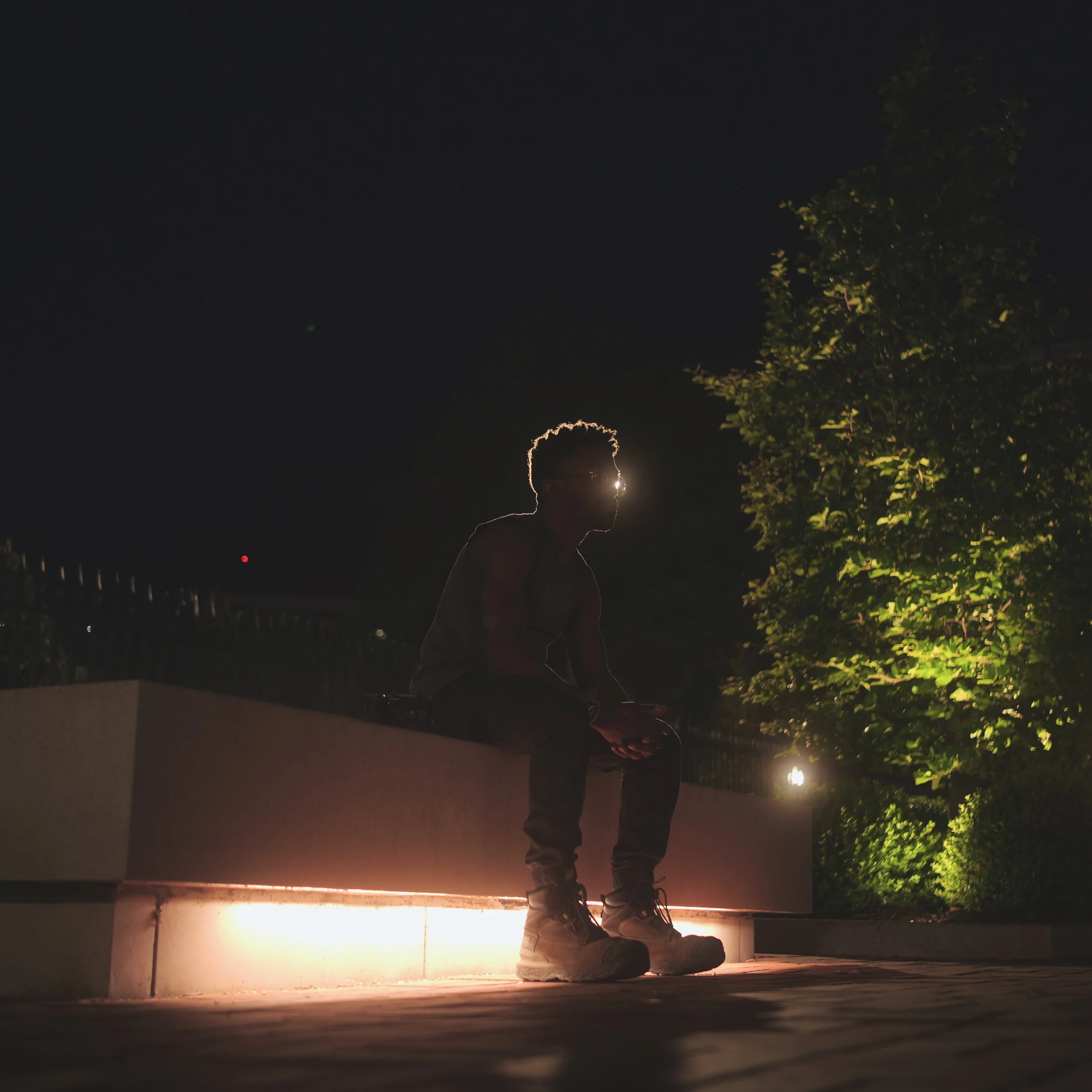 A person standing outdoors at night, wearing sneakers, with trees visible on the right side, and a bright light source behind them illuminating their silhouette.