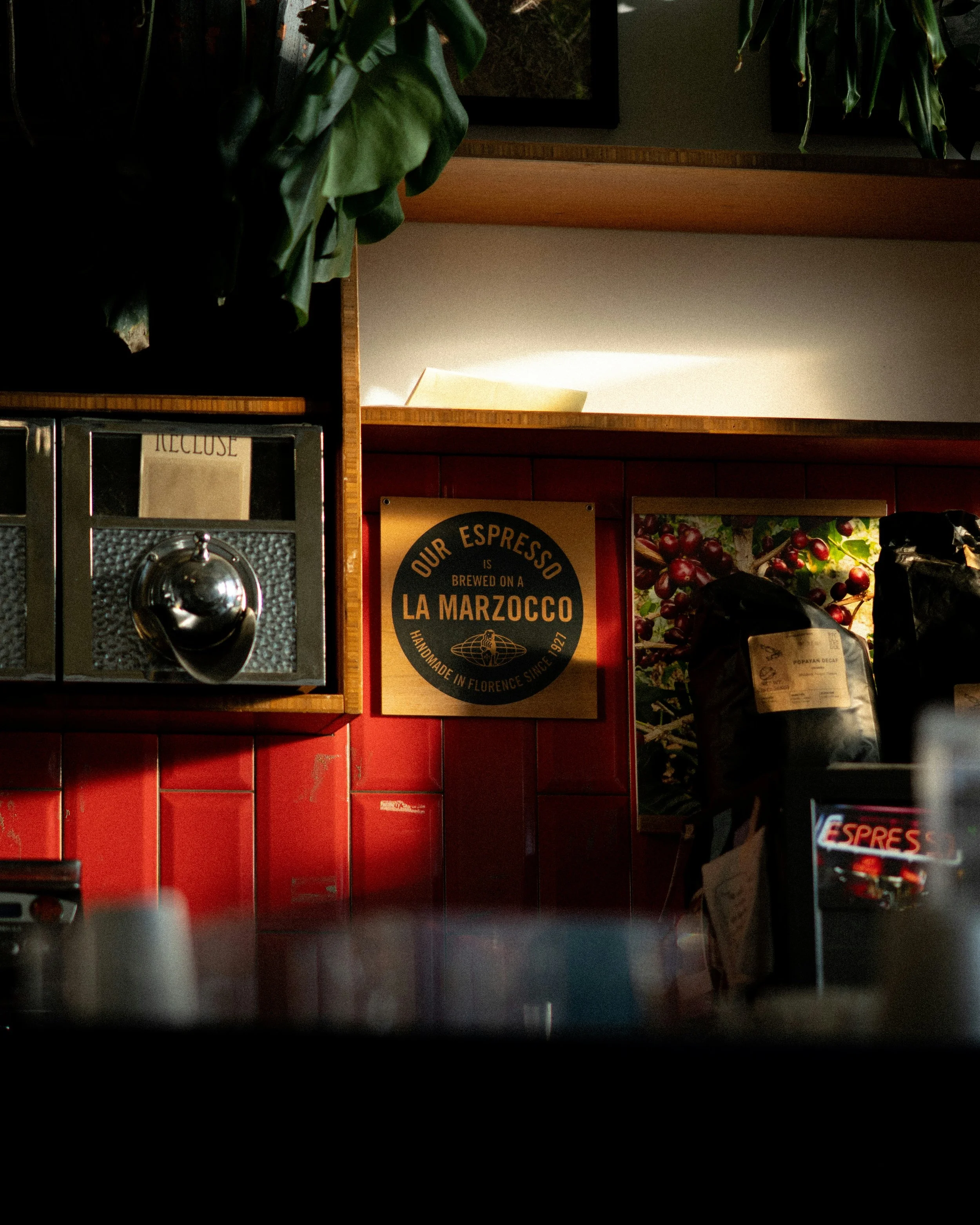 The image shows a coffee shop setting with red tiles on the lower wall, a sign indicating that their espresso is brewed on a La Marzocco machine, a poster displaying red cherries, and part of a black espresso machine with a metallic knob at the botto
