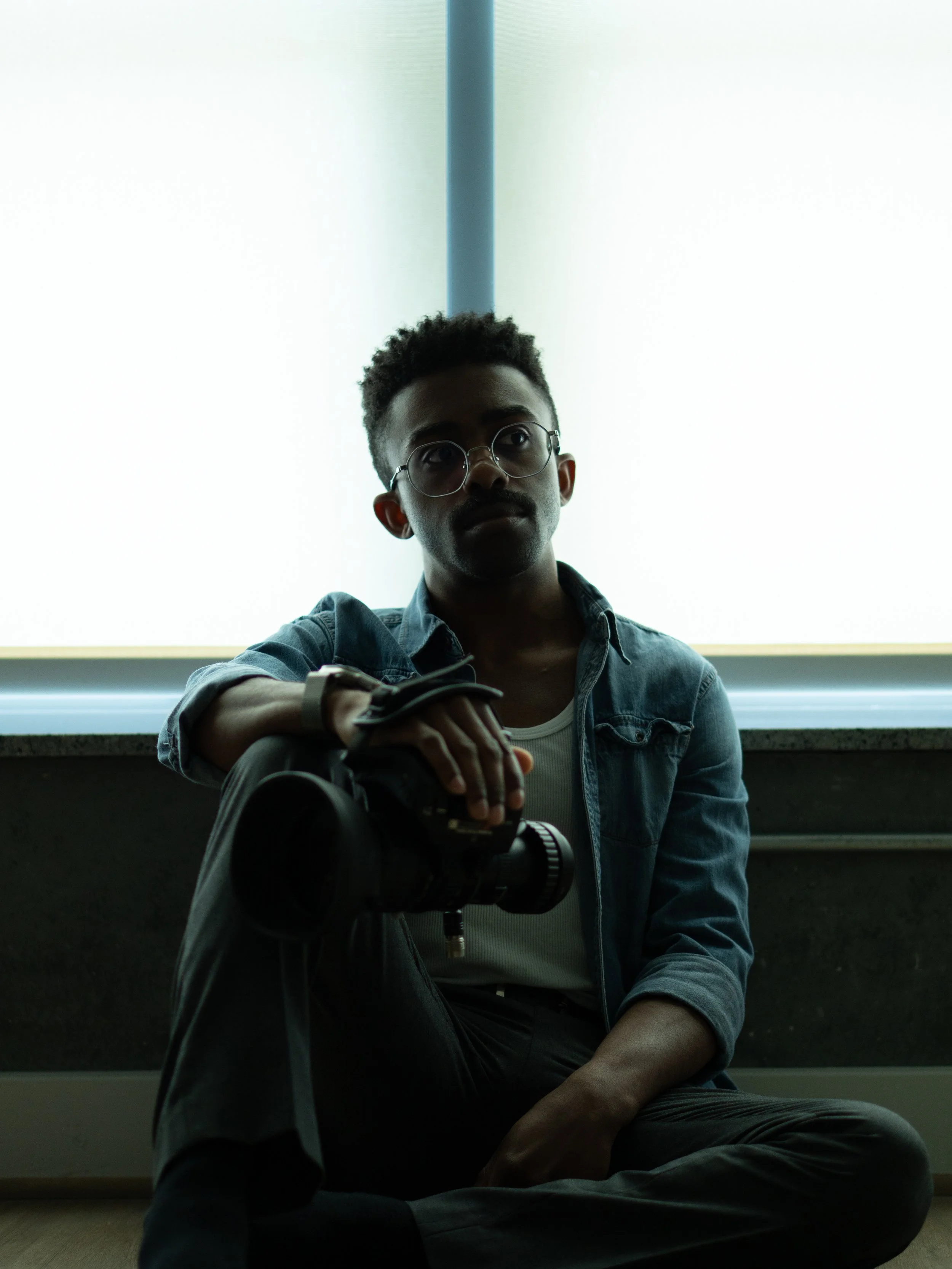 A young man with glasses and a denim jacket sitting on the floor by a large window, holding a camera with a strap, in a room with natural light.