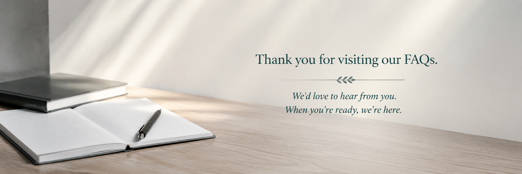 A thank you message on a wall that reads, 'Thank you for visiting our FAQs. We'd love to hear from you. When you're ready, we're here.' next to a wooden desk with an open notebook, a pen, and a closed book.