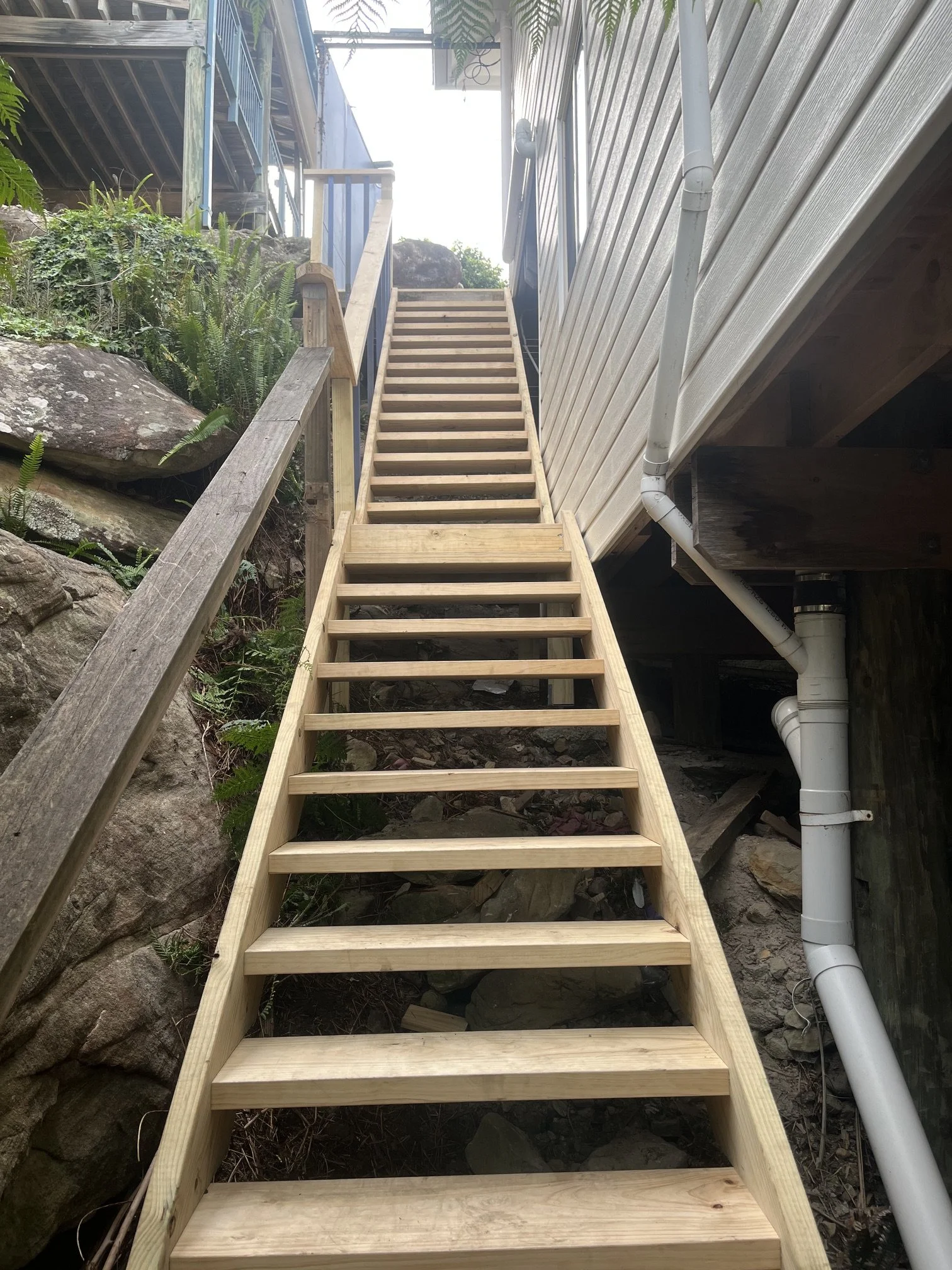 Wooden stairs leading upwards next to a house with piping underneath, surrounded by rocks and plants.